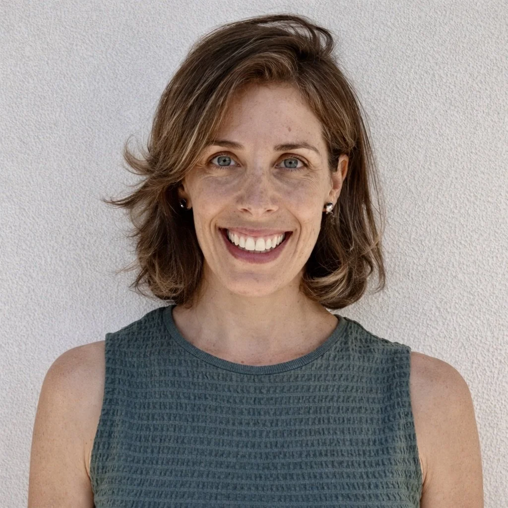 A woman with short, wavy brown hair, light blue eyes, and fair skin, smiling and standing against a plain white wall, wearing a sleeveless teal textured top.