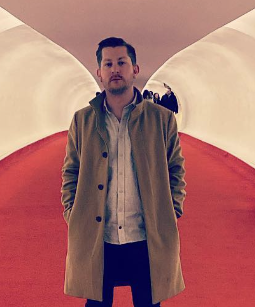 A man standing in an abstract indoor space with curved white and red walls, with a small group of people visible in the background.
