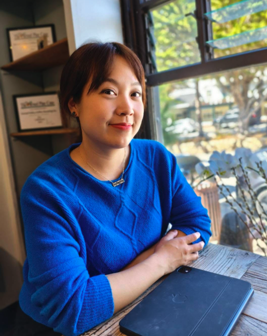 A woman sitting at a wooden table in a cafe, looking at the camera and smiling, with a laptop in front of her and sunlight coming through a window behind her.