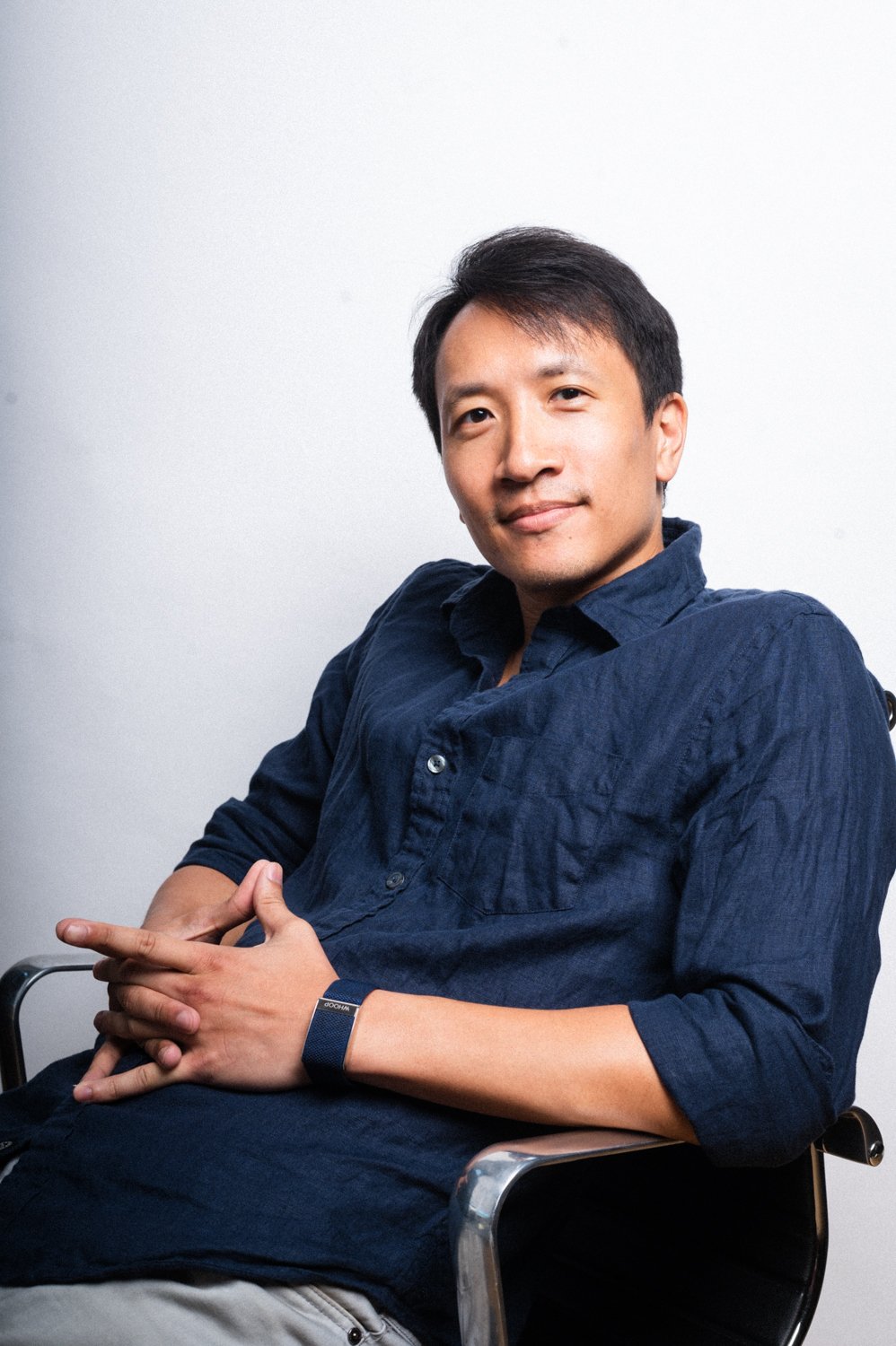 A young man sitting in a black office chair, wearing a navy blue button-up shirt, with arms crossed and hands clasped, against a plain white wall background.