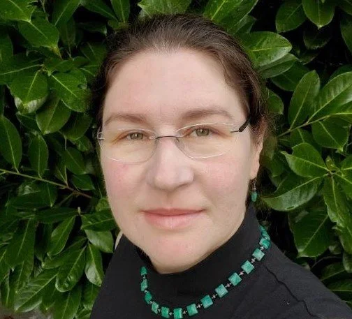 Portrait of a woman with glasses, dark hair, and a necklace, standing in front of green leafy plants.