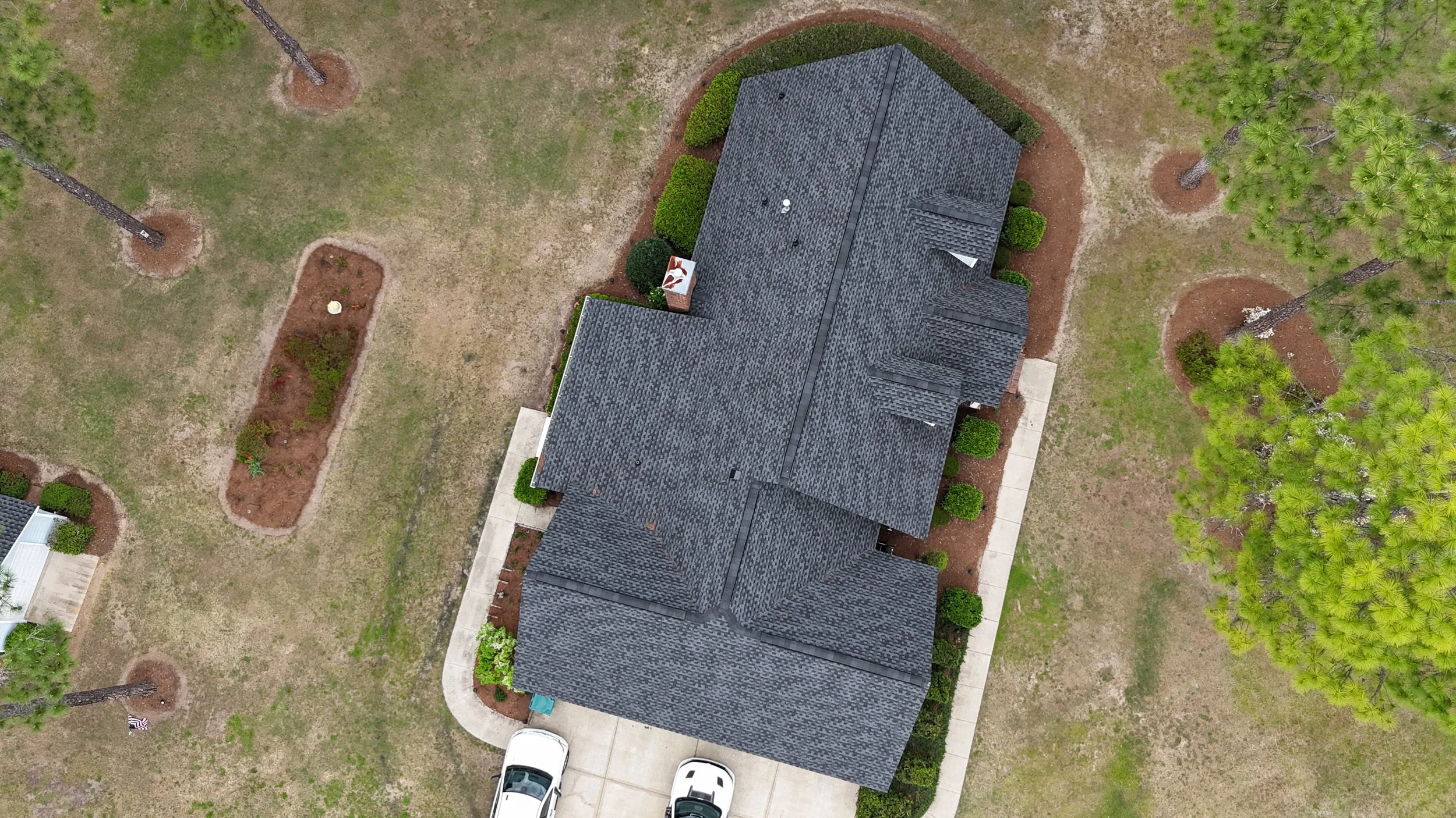 Aerial view of a house with a dark gray roof, surrounded by a landscaped yard with trees, bushes, and a concrete pathway, with two parked cars in the driveway.