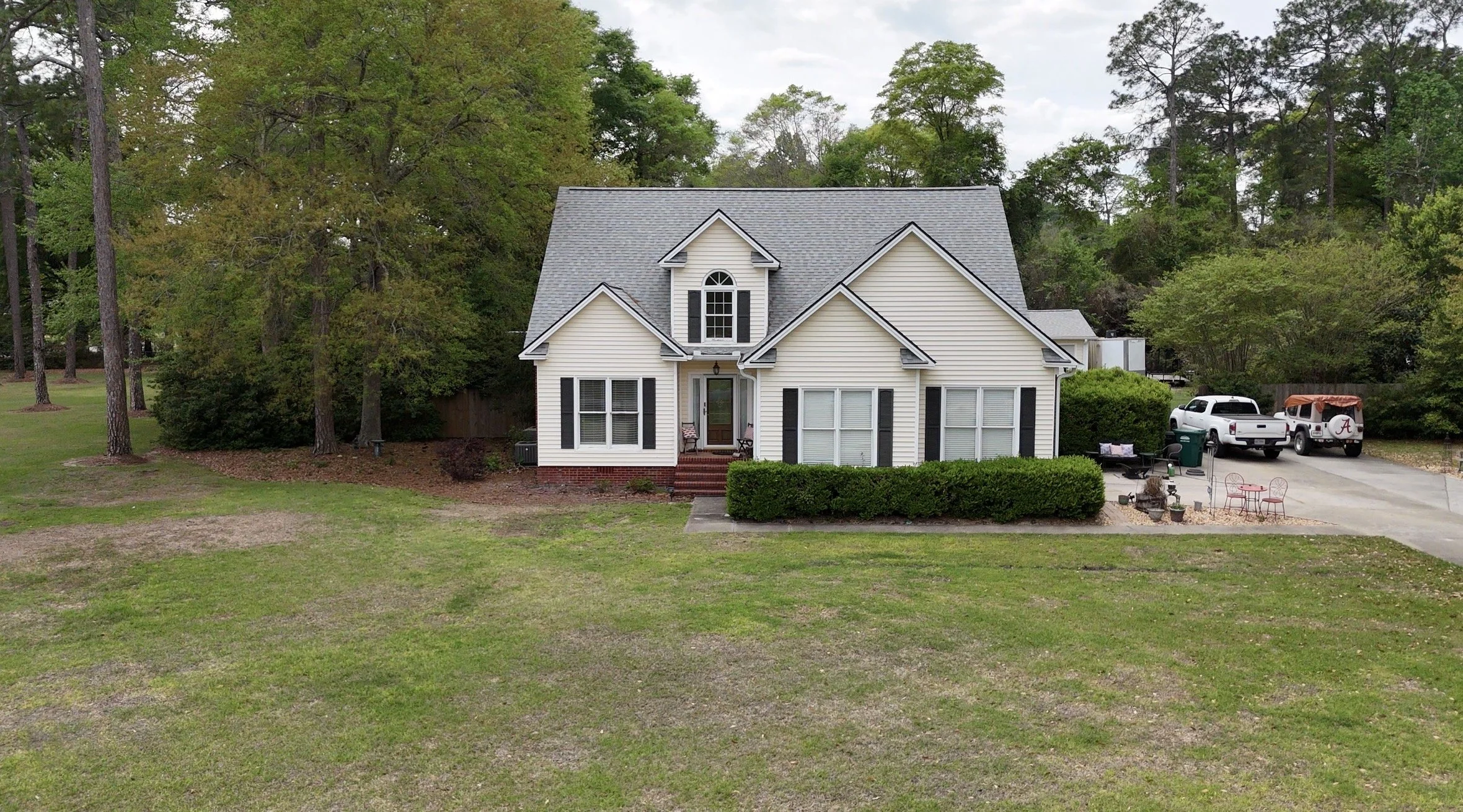 A two-story white house with black shutters and a gray roof, surrounded by green trees and a lawn, with a driveway on the right side leading to parked vehicles and outdoor seating area.