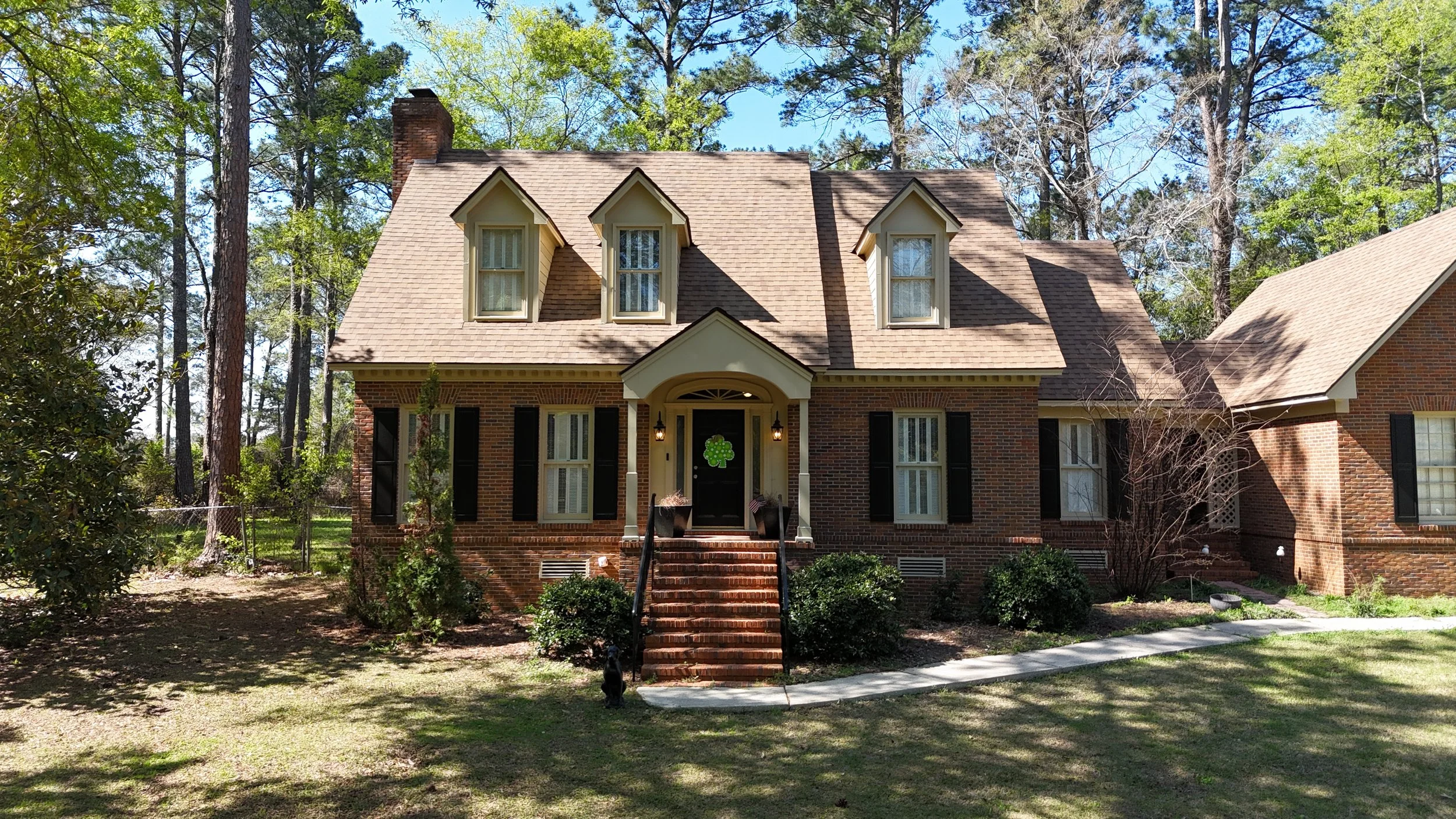 A brick house with a brown shingle roof, three dormer windows, black shutters, and a small front porch with stairs, surrounded by trees and greenery.