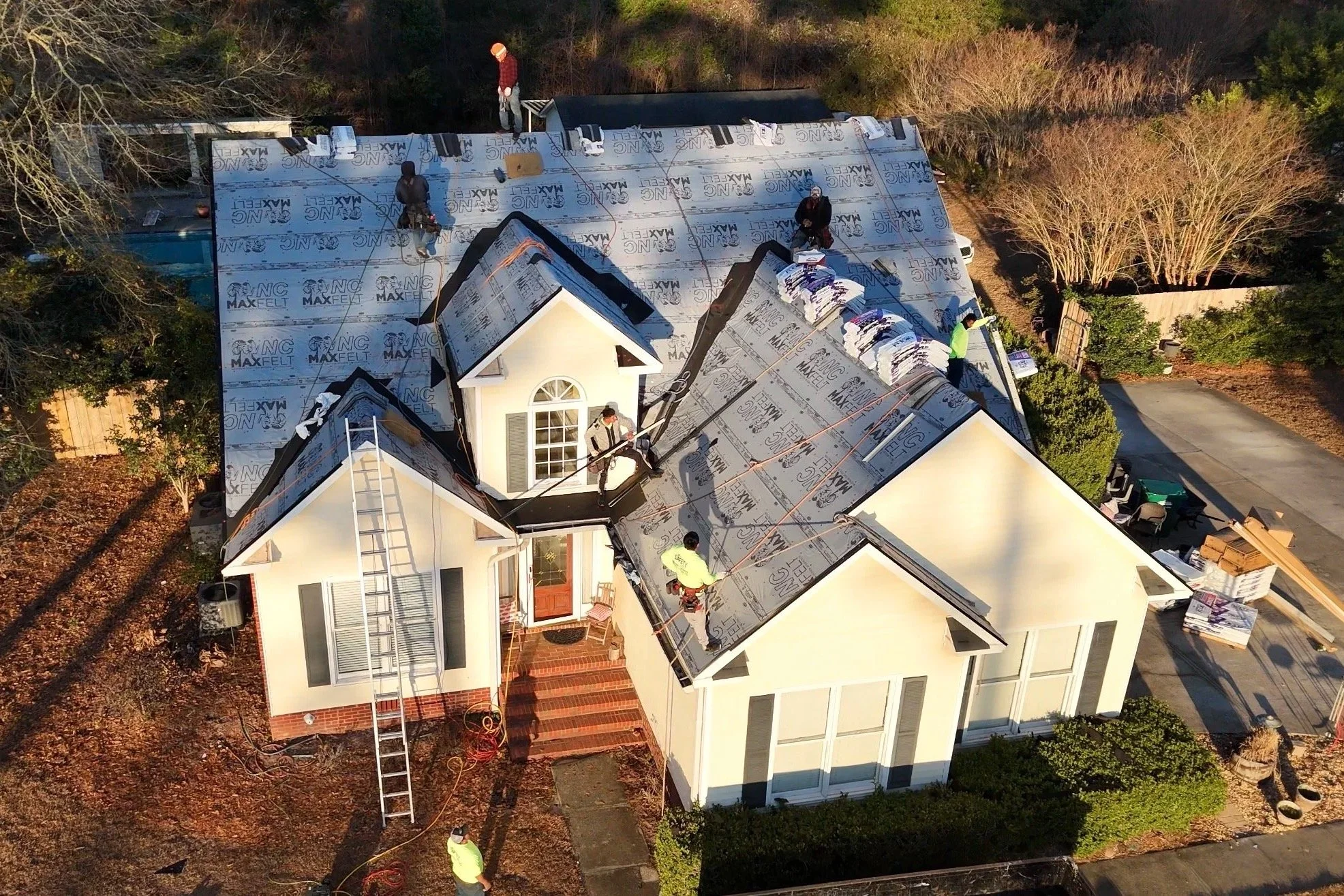 An aerial view of a house under construction with workers installing a new roof, which is covered with roofing materials and underlayment. The house features a white exterior with a front porch and a tall ladder leaning against it. Several workers are on the roof, and others are on the ground around the house.
