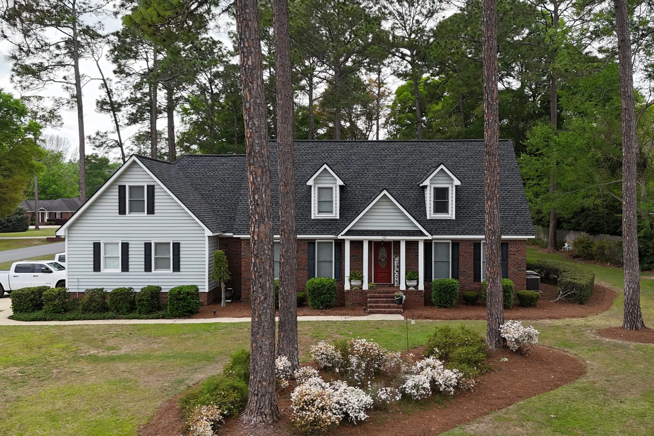 A two-story house with a brick and white siding exterior, black shutters, and a red front door surrounded by tall pine trees, with landscaped bushes and white flowering plants in the front yard.