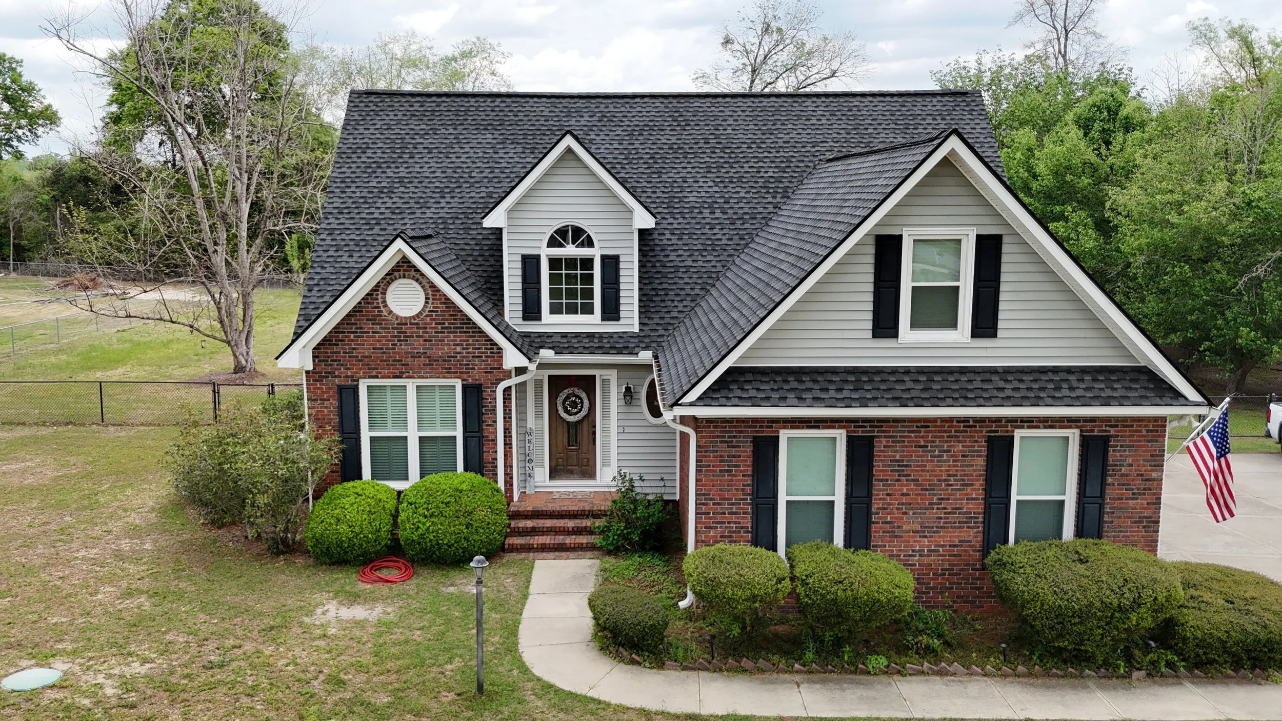 Front view of a two-story house with red brick and white siding, black shutters, a black shingled roof, and a front porch with steps, surrounded by bushes and a lawn, with American flag on the right side.