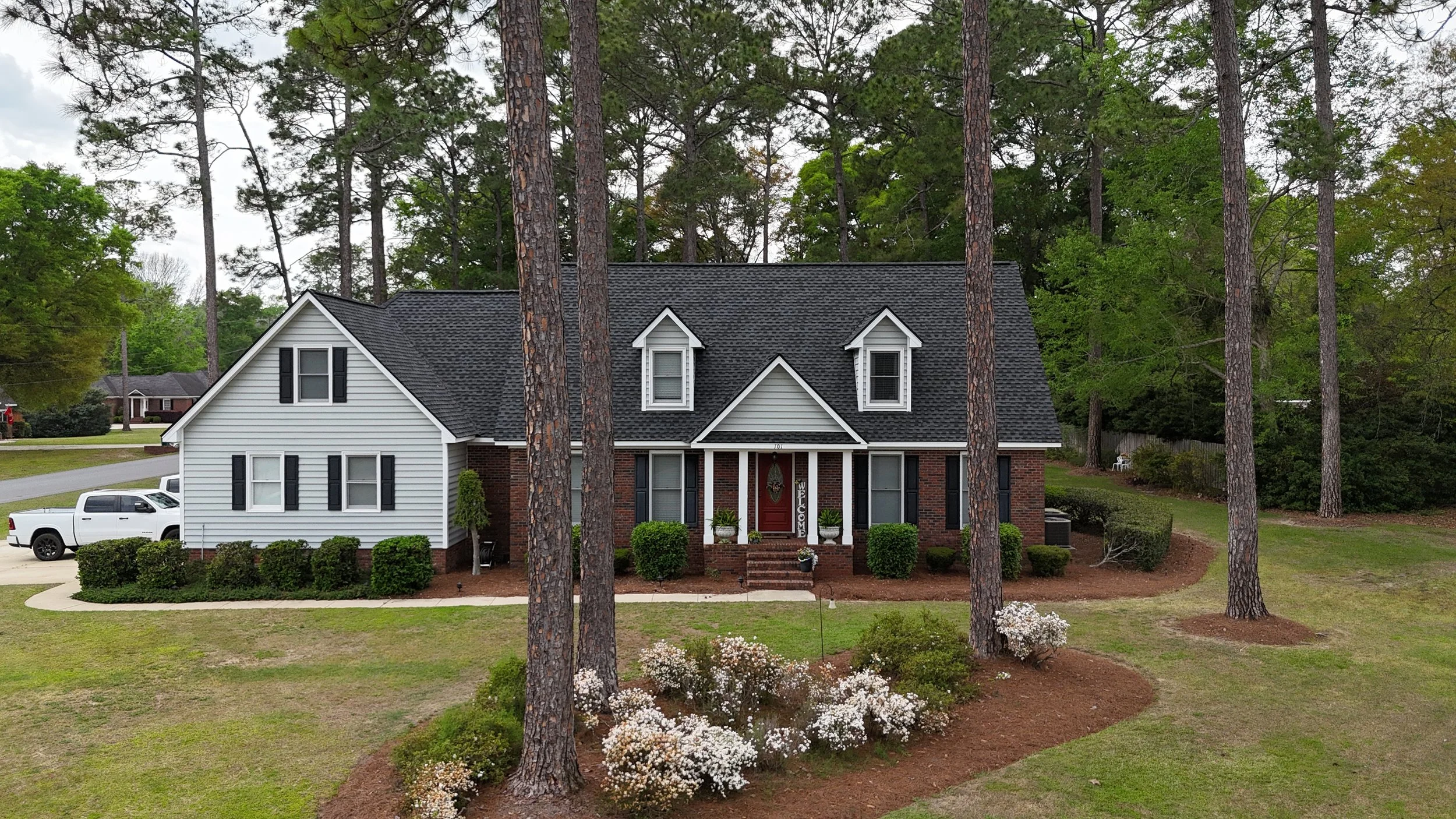 Front view of a two-story house with brick and white siding, black shutters, a dark shingled roof, and a welcoming front porch with steps, surrounded by tall pine trees and landscaped gardens with bushes and white flowers.