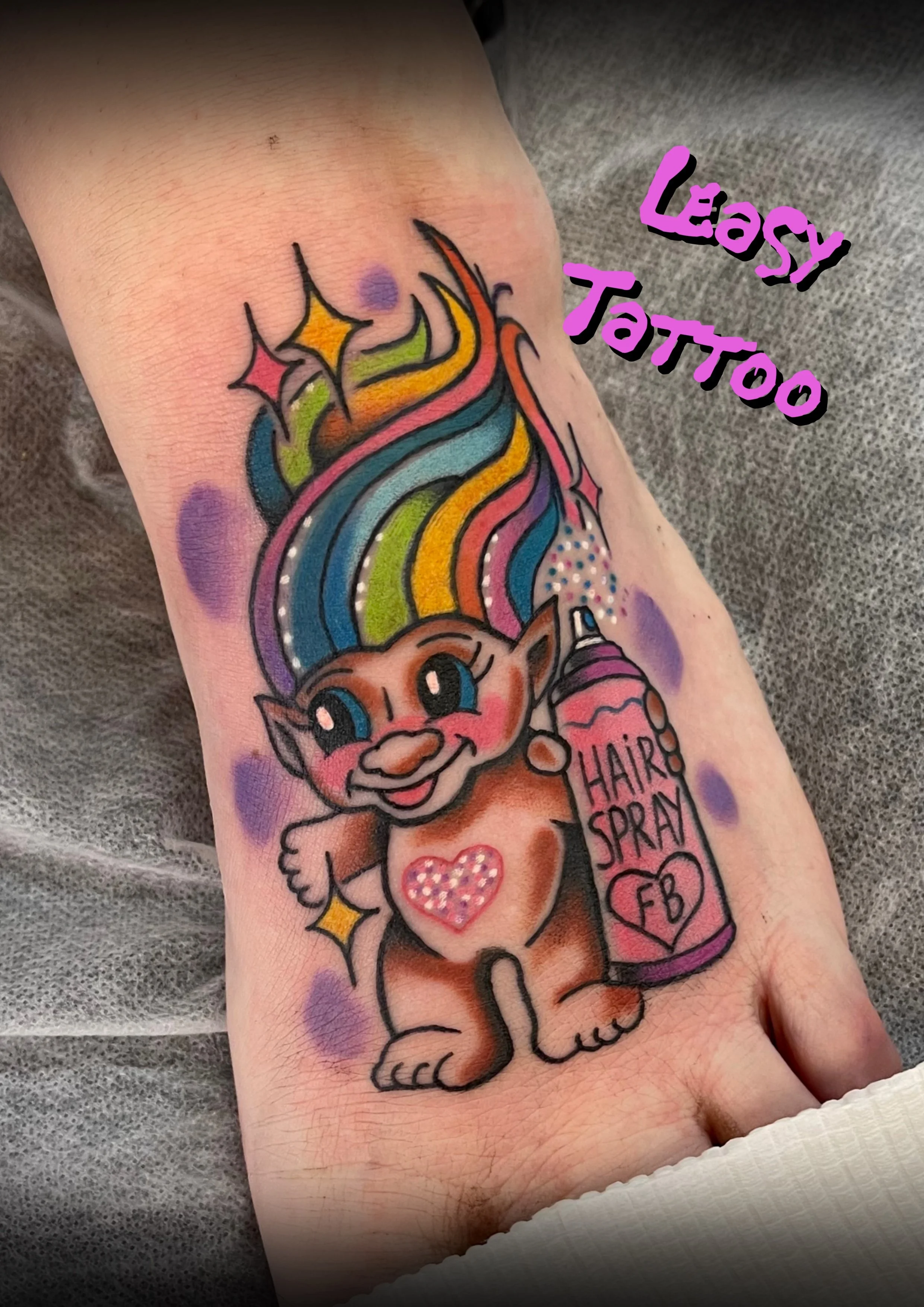 Colorful tattoo of a unicorn with a rainbow mane and tail, holding a heart-shaped spray can labeled 'Hair Spray.' The tattoo has sparkles and stars, with text nearby reading 'Lazy Tattoo.'