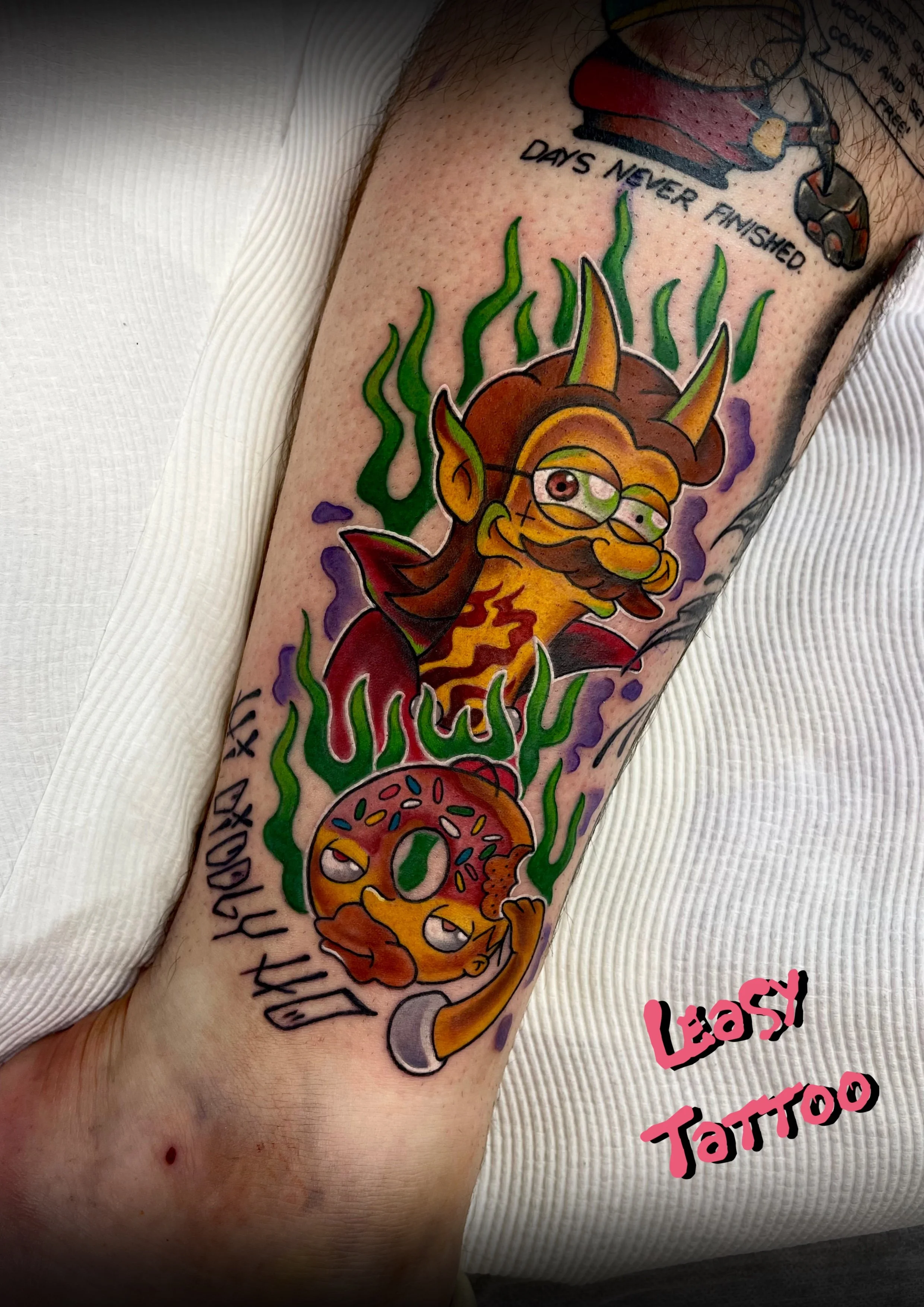 Colorful tattoo on person's arm featuring a cartoonish devil character with horns, wings, and a mischievous expression, surrounded by green flames and purple smoke. Text includes 'DAYS NEVER FINISHED' and 'LEASH TATTOO'.
