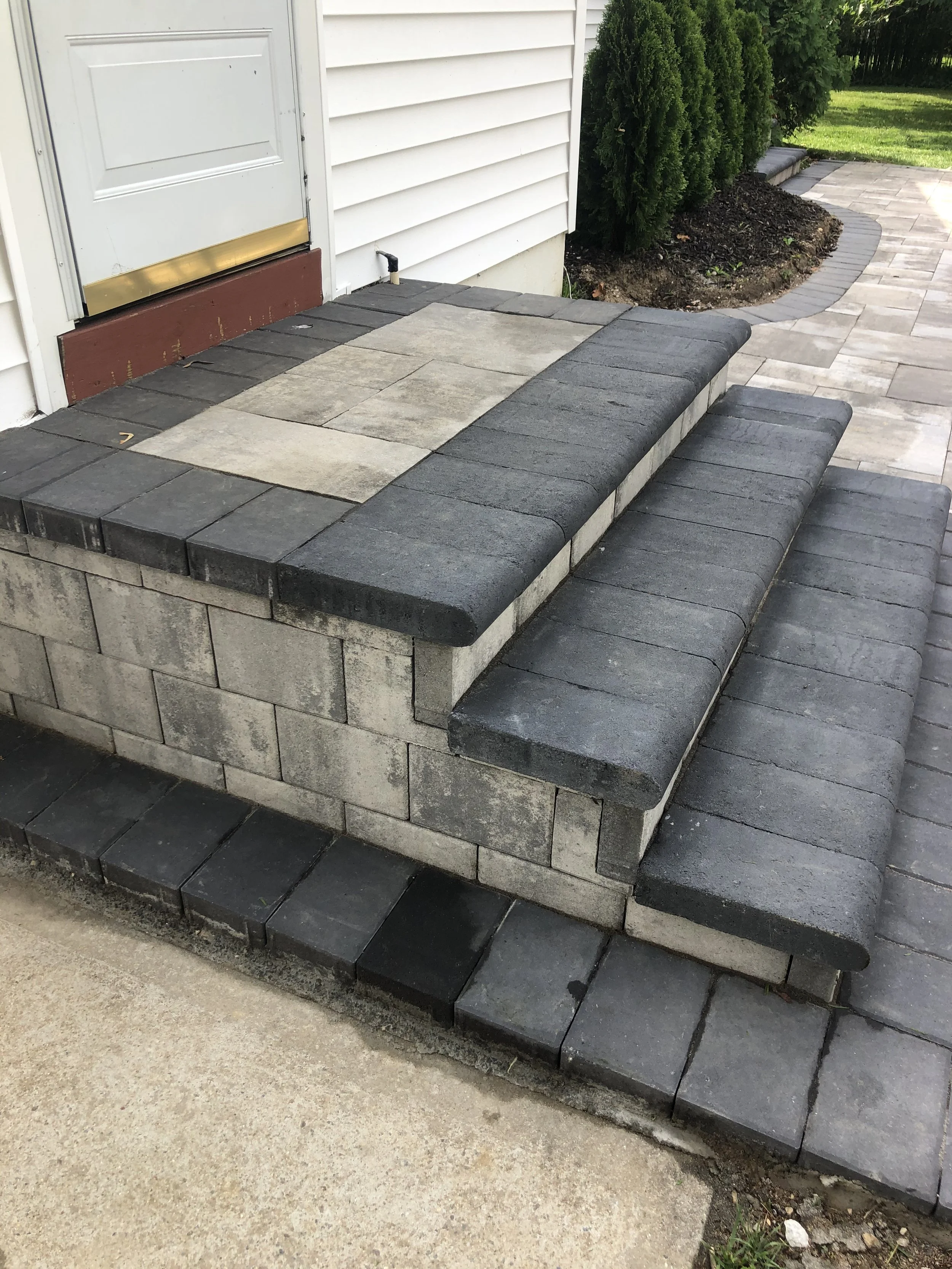 A raised porch with stacked concrete and stone pavers, with a small set of steps leading up to a white door, surrounded by shrubbery and a paved walkway.