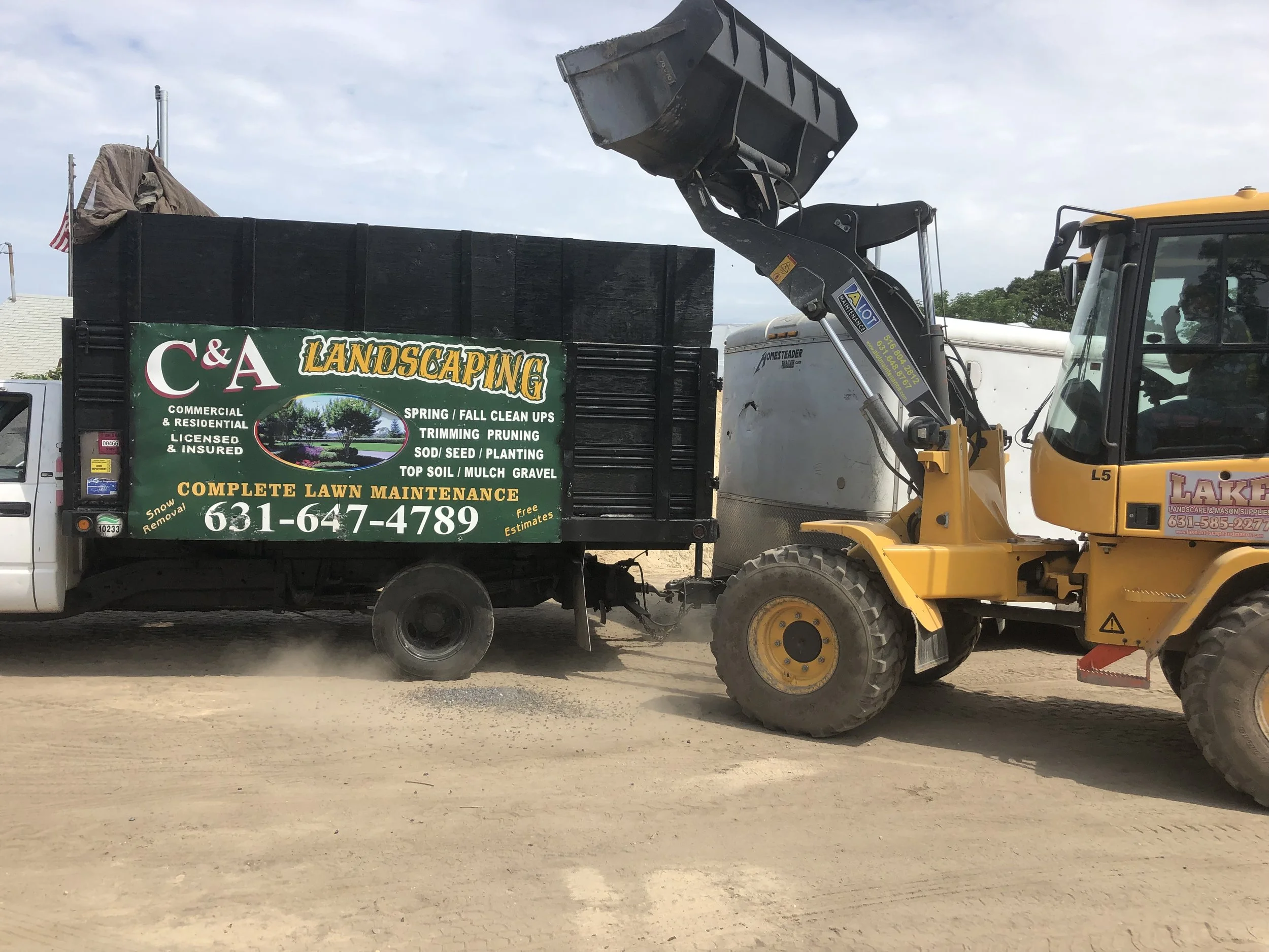 A front loader is attaching to a black truck with a green sign that reads "C&A Landscaping" offering lawn maintenance services, in a dirt area with a partly cloudy sky.