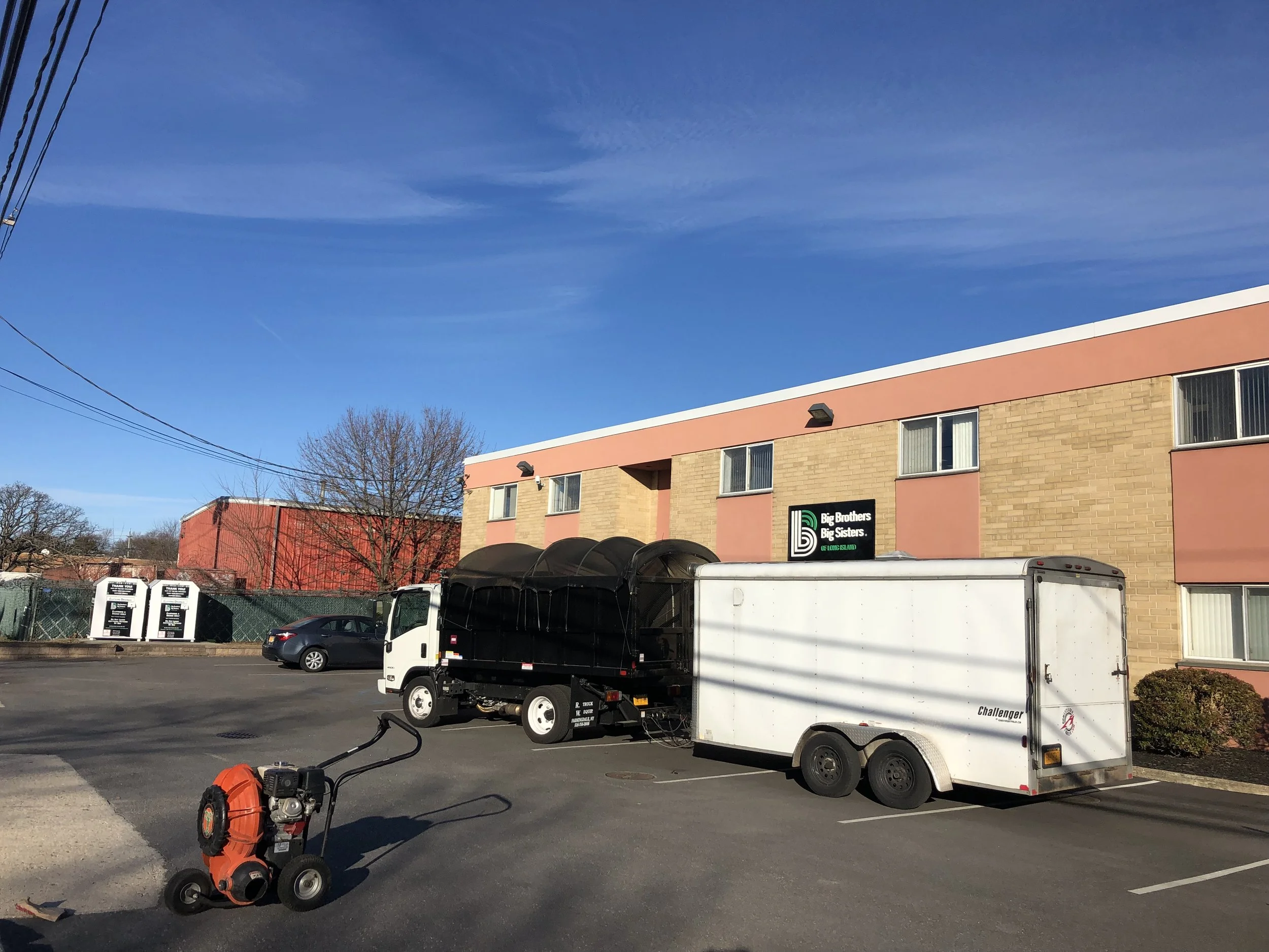 Parking lot with a large black and white truck and trailer, a small orange and black pressure washer, and a two-story beige and pink building with a sign that reads "Big Brothers Big Sisters" under a partly cloudy blue sky.