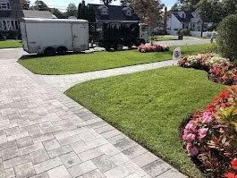 A well-maintained lawn with pink flowers along the edge, a paved walkway, and trees in the background.