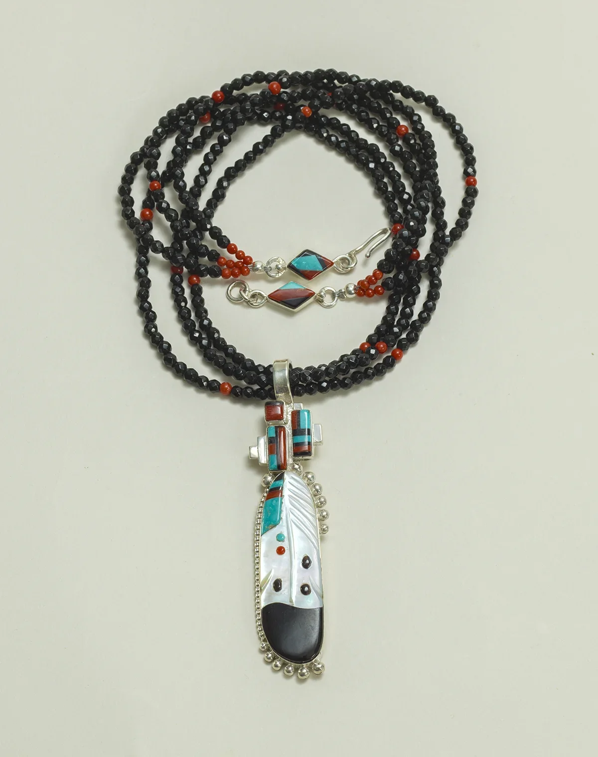 Two sets of black beaded necklaces with small red beads, adorned with silver and turquoise jewelry, including a feather pendant and geometric charms.