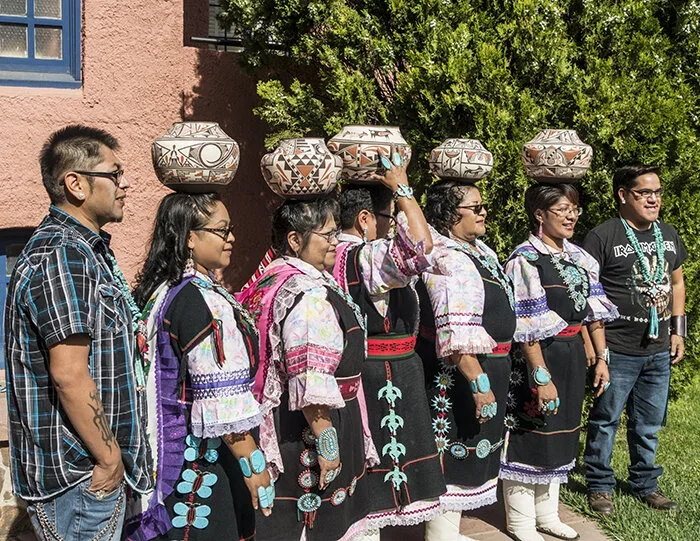 Group of seven women and two men standing outdoors, dressed in traditional Zuni clothing, some balancing large decorated pottery on their heads, with green bushes and a pink stucco building in the background.