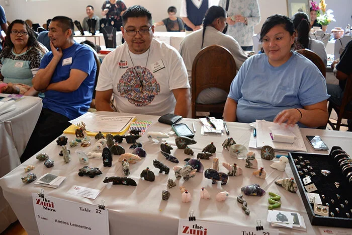 People sitting at a table displaying various decorative rocks, gemstones, and jewelry items at the Zuni Show