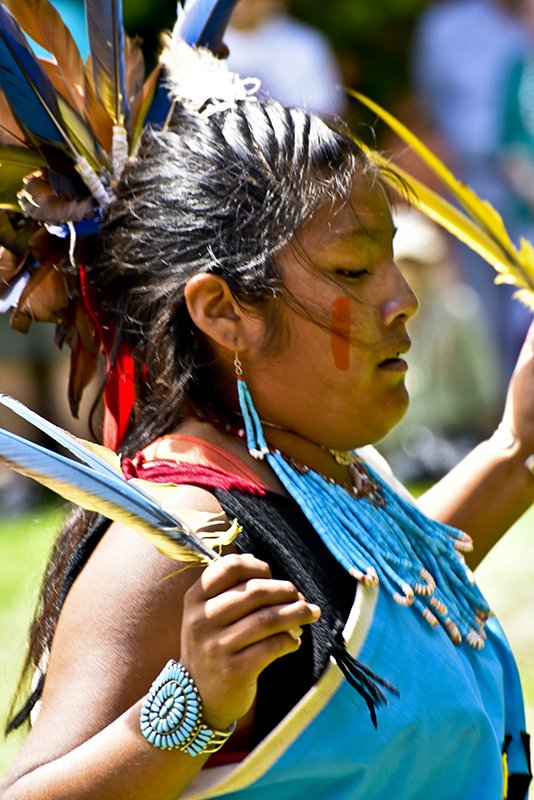 Young Indigenous woman wearing traditional Native American attire, including a feather headdress, colorful beaded jewelry, and face paint, participating in a cultural ceremony at the Zuni Show.