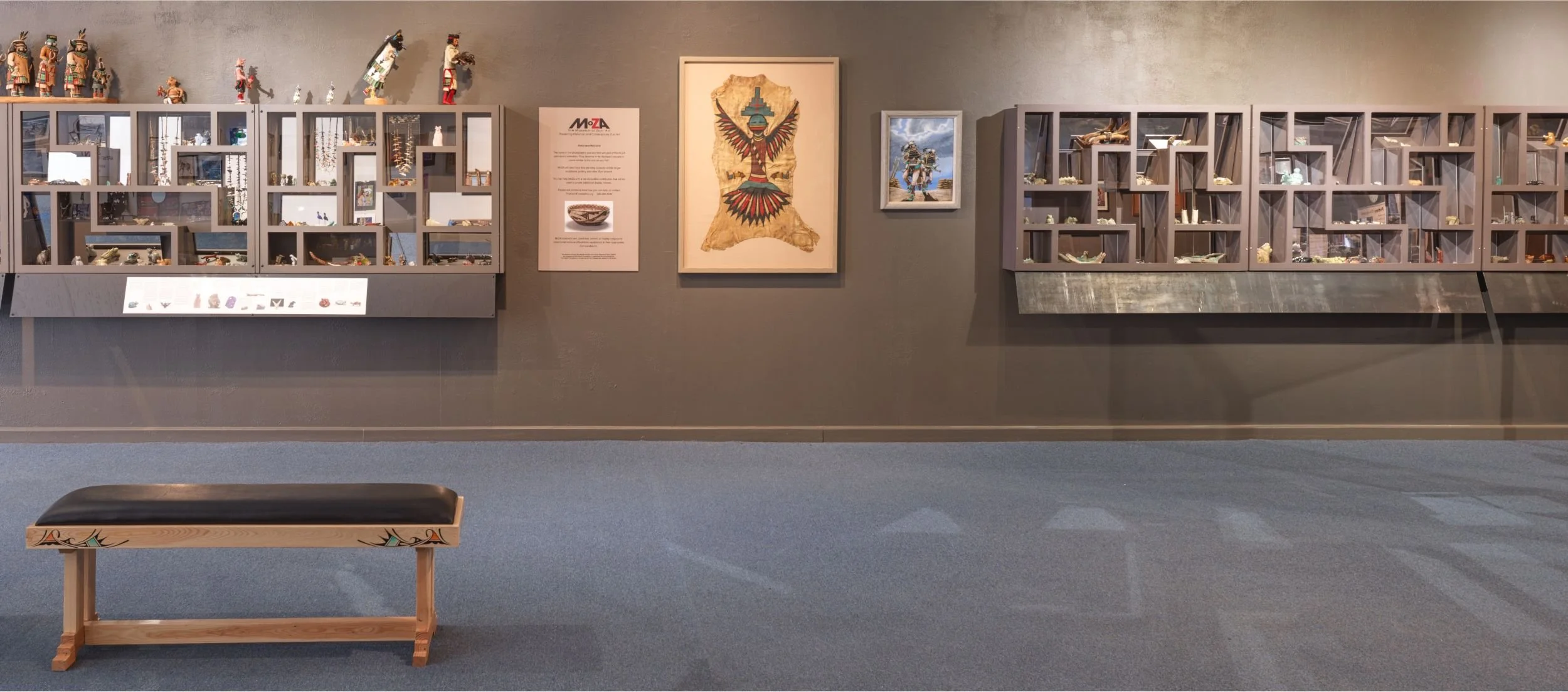A museum of Zuni Art MoZA display with glass cases containing Zuniartifacts, framed artwork on the wall, and a wooden bench in front.