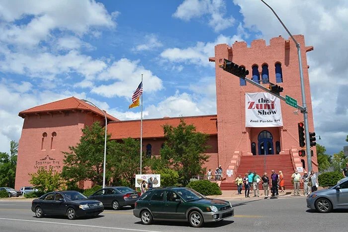 Santa Fe Scottish Rite building, featuring a banner for the Zuni Pueblo Art Show, American flag and trees in front, several people walking nearby, and cars parked in front.