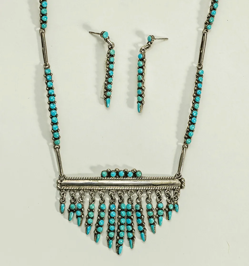 Joyce Eriacho Snake eyes. A set of jewelry including a necklace and a pair of earrings, all with turquoise stones and silver metal, arranged on a white surface.