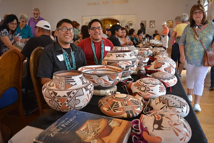 Two young men sitting behind a table filled with various native pottery and art, smiling at the camera during a cultural event in a hall with many people in the background.