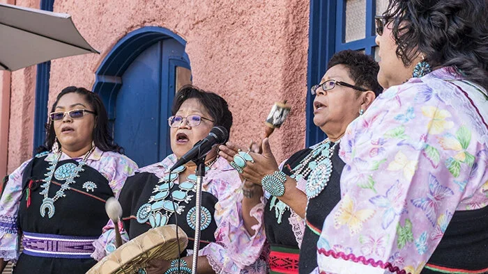 Four women wearing traditional Mexican clothing and jewelry singing karaoke outdoors, standing in front of a pink stucco wall with a blue door.