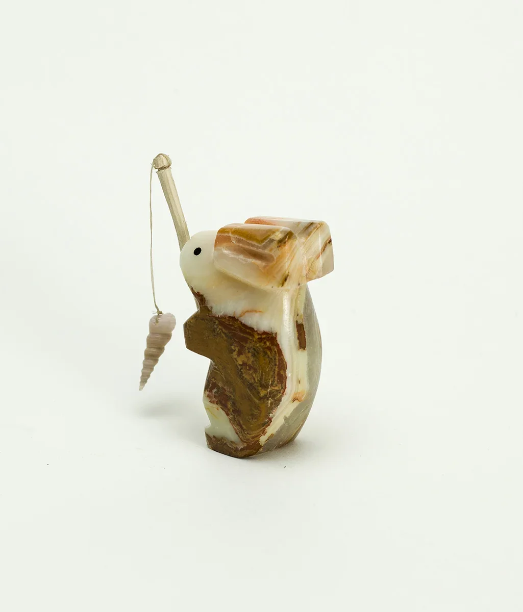 A small sculpture of a rabbit made from a shell and stone, holding a fishing rod with a tiny fish attached. The sculpture is minimalist with a smooth, polished surface, and is set against a plain white background.