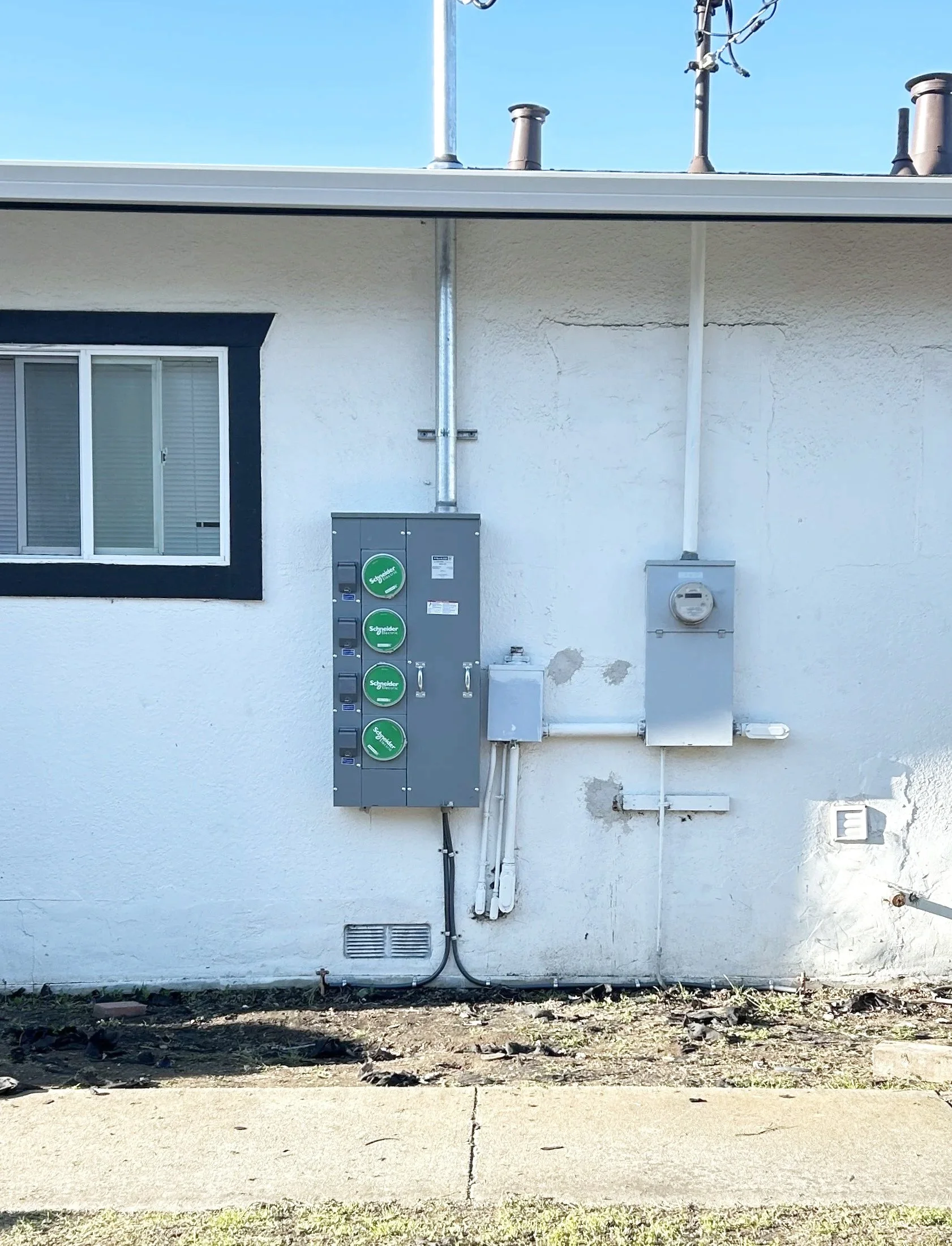 Exterior side of a house with a window, utility boxes, pipes, and electrical meters attached to the white wall.
