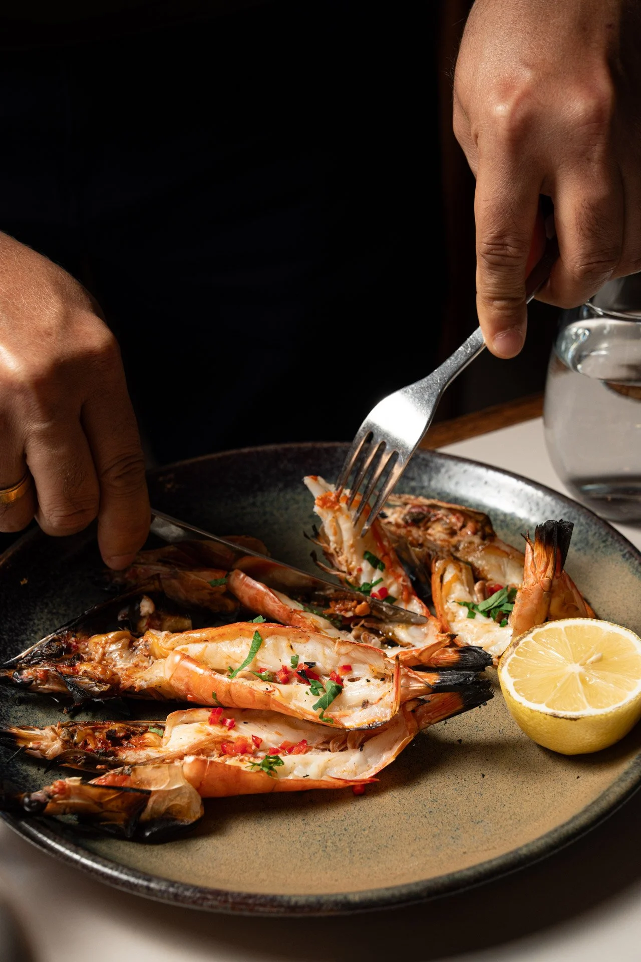 Albury greek best restaurant: Person using a fork to eat grilled shrimp garnished with herbs and red pepper flakes, served with a lemon half on a dark plate.
