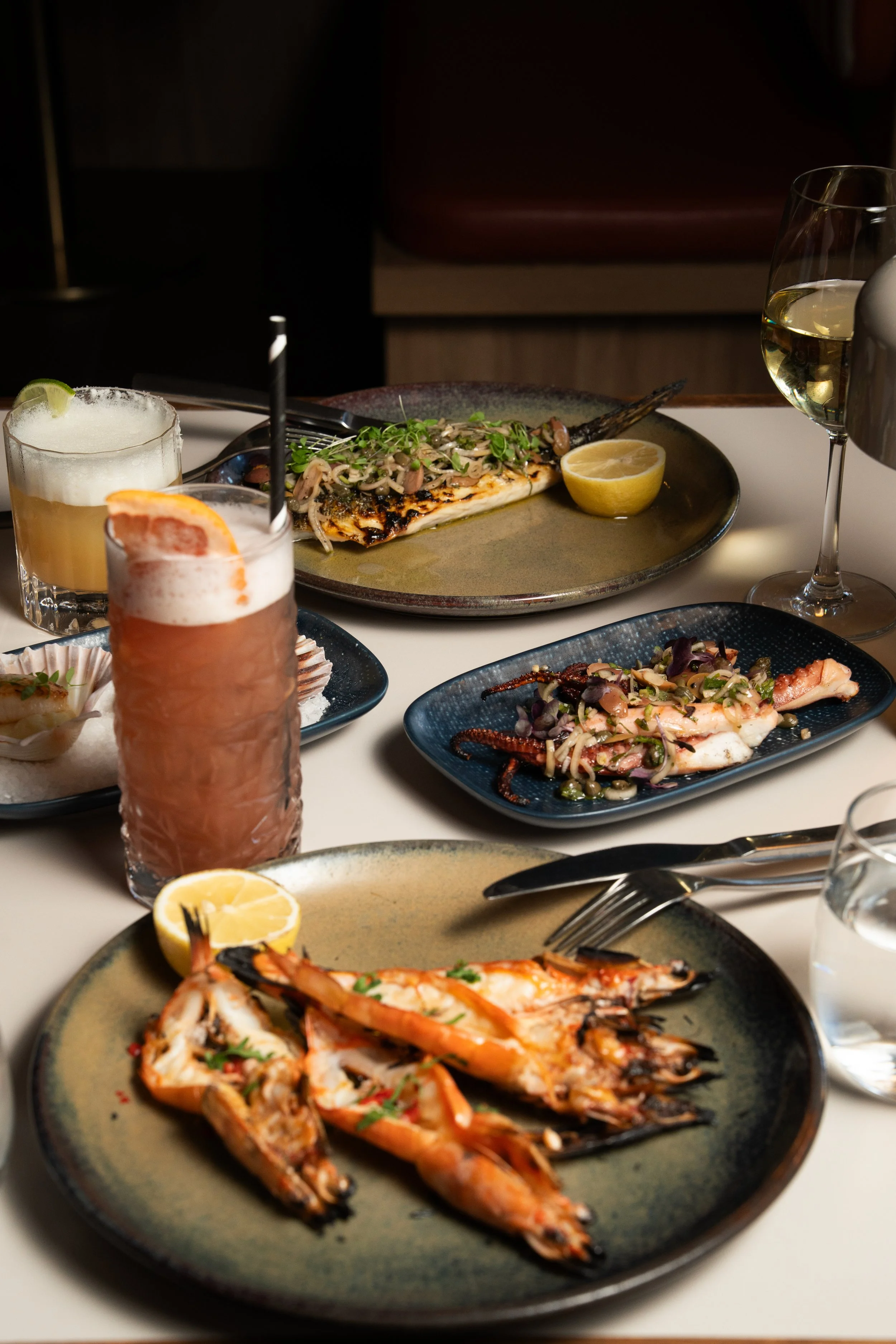 Albury greek best restaurant: A seafood dinner including grilled shrimp, fish topped with herbs and seafood, and a seafood salad, with drinks such as wine, cocktails, and water on a restaurant table.