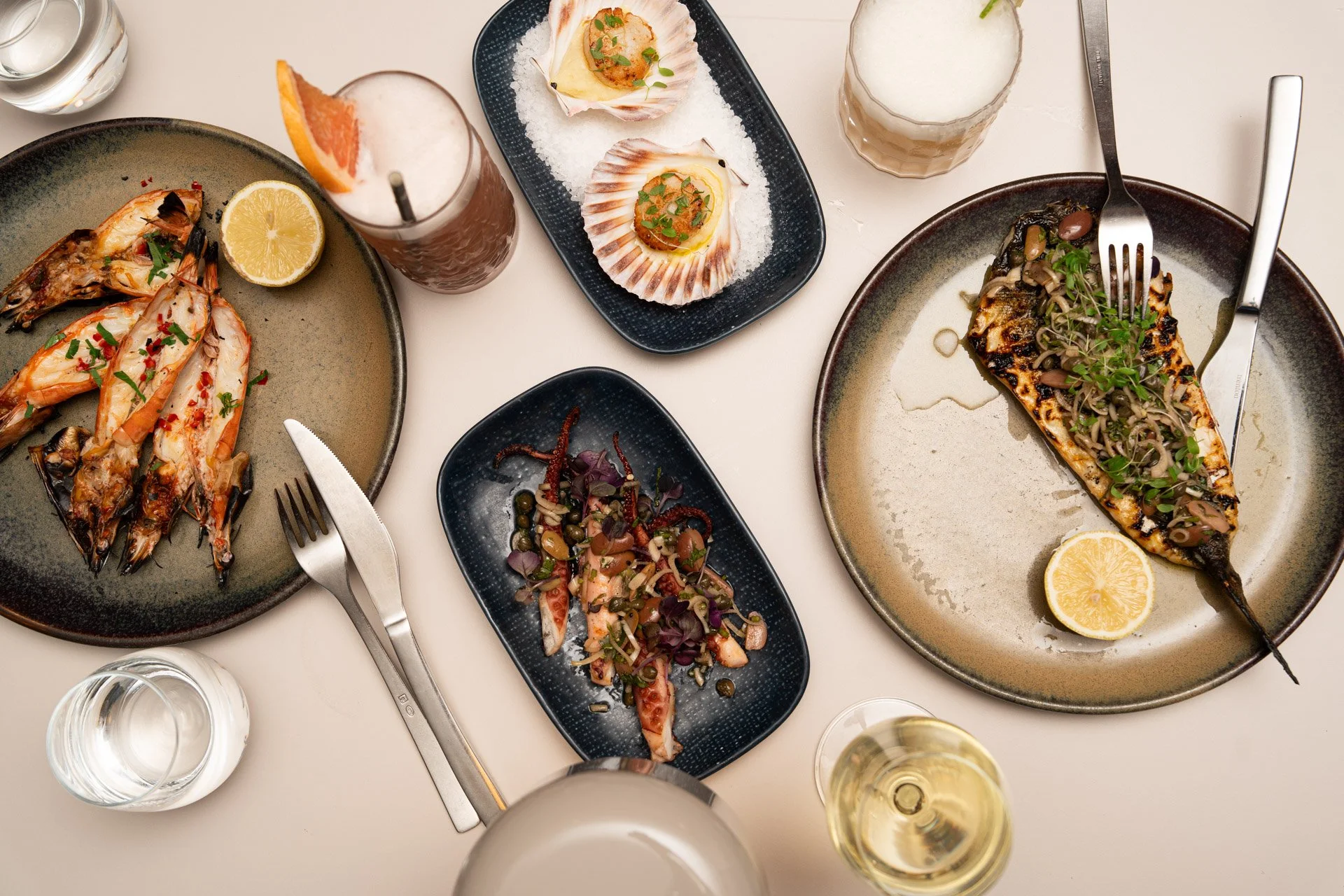 Albury greek best restaurant:  A table with various seafood dishes, including grilled fish with herbs, shrimp, a seafood salad, and scallops, along with drinks like beer, white wine, and cocktails.