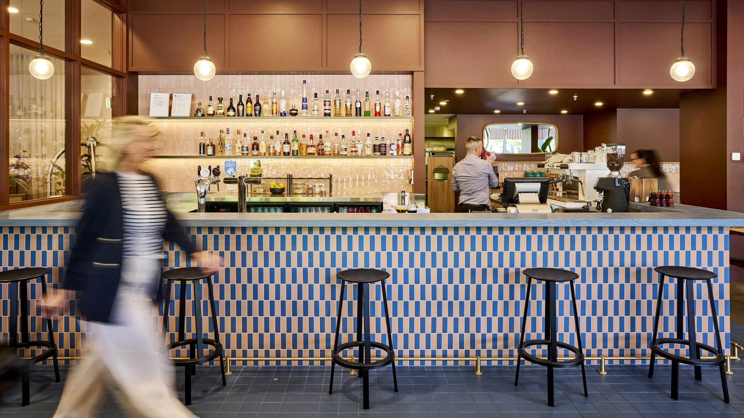 Albury greek best restaurant: Modern bar with colorful geometric tiled front, bottles of alcohol on shelves, bartender preparing drinks, customer standing at the bar, and a blurred woman walking past.