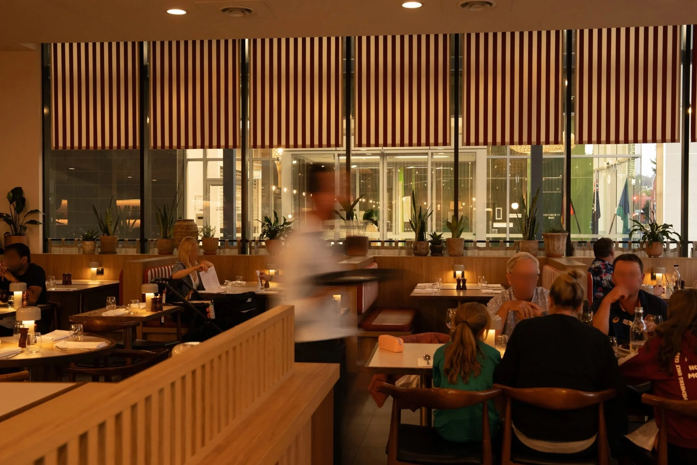 Albury greek best restaurant: Interior of a restaurant with wooden furniture, potted plants, and diners seated at tables. Large windows with striped blinds are in the background, and a blurred waiter is walking through the scene.