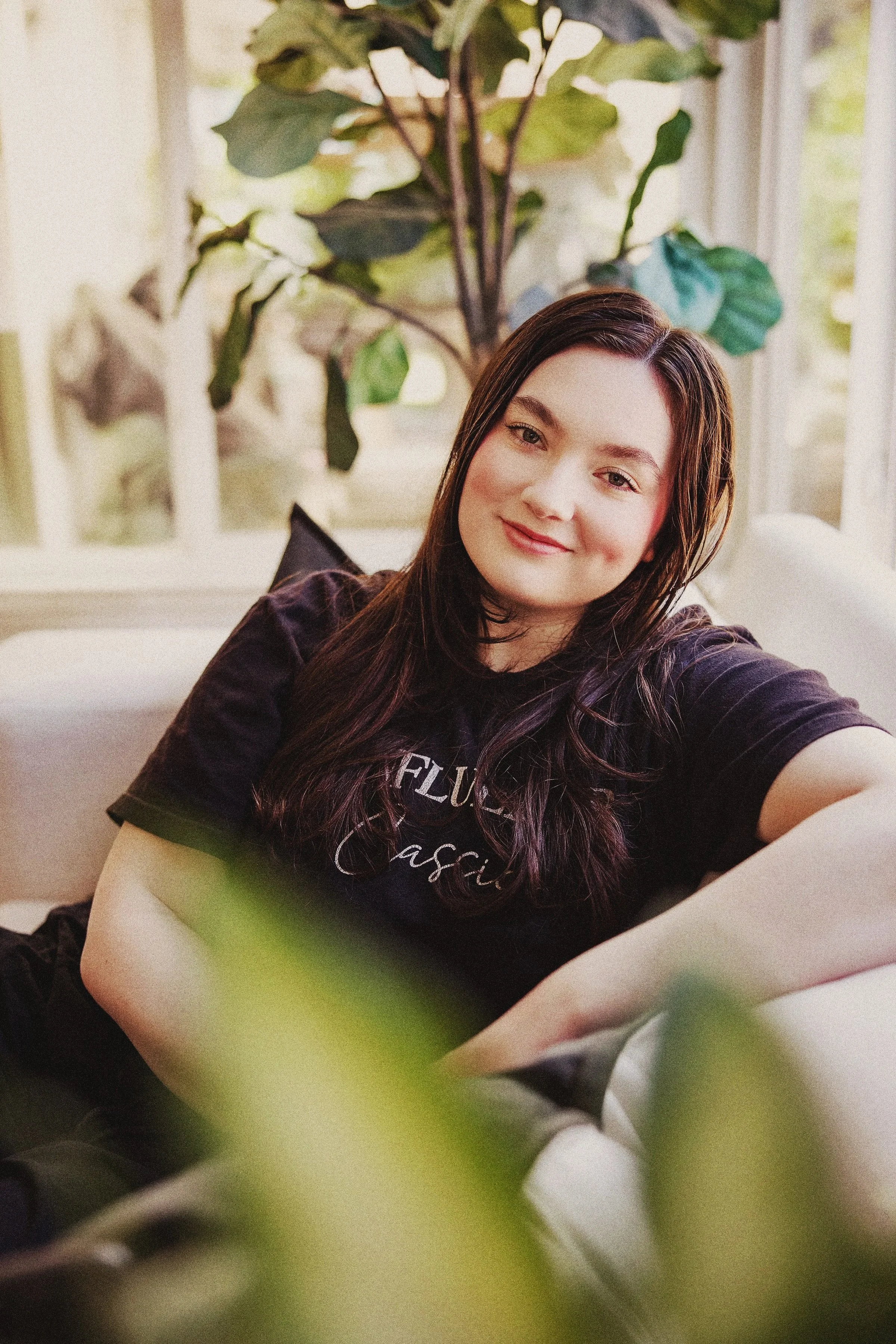 A young woman with long dark hair, smiling, taking a selfie in a bright room with large windows and a green plant in the background.