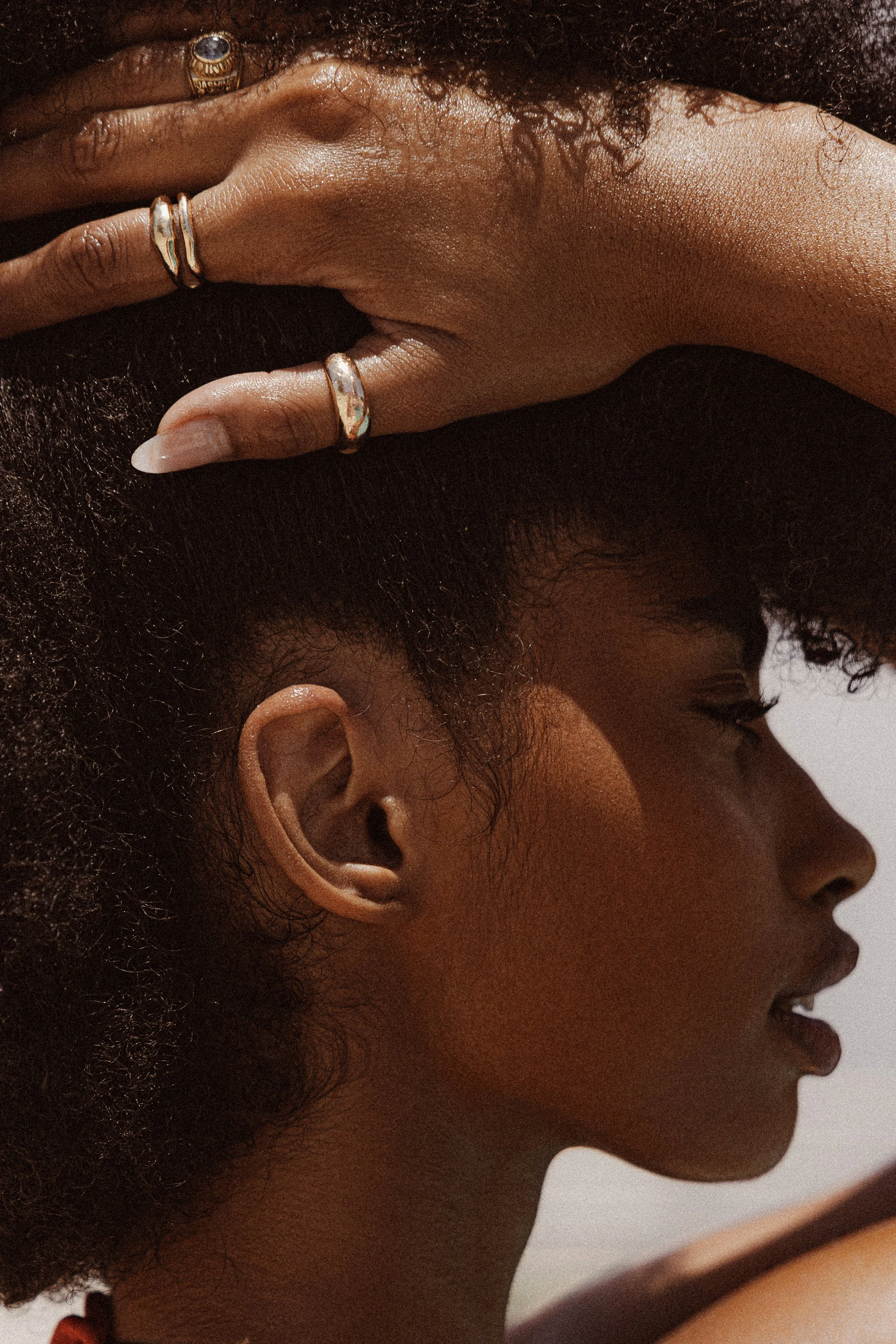Close-up of a woman's face and hand, showing curly hair, earrings, and rings, with a neutral background.