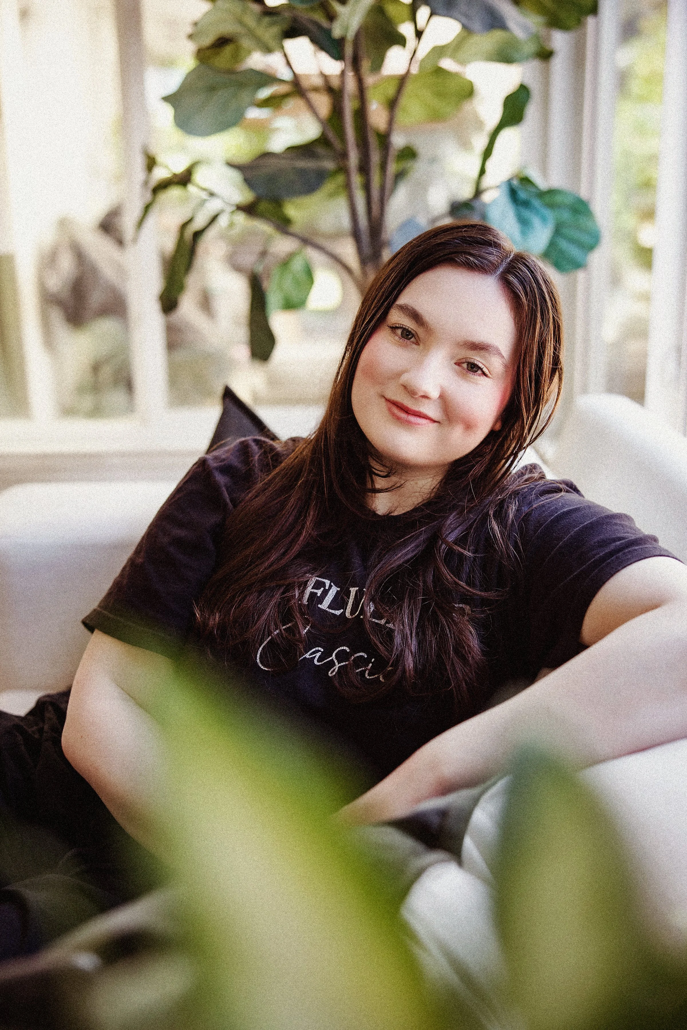 A young woman with long, dark brown hair and fair skin sitting on a green chair, smiling softly at the camera. Behind her, there is a large potted plant with broad green leaves near a window.