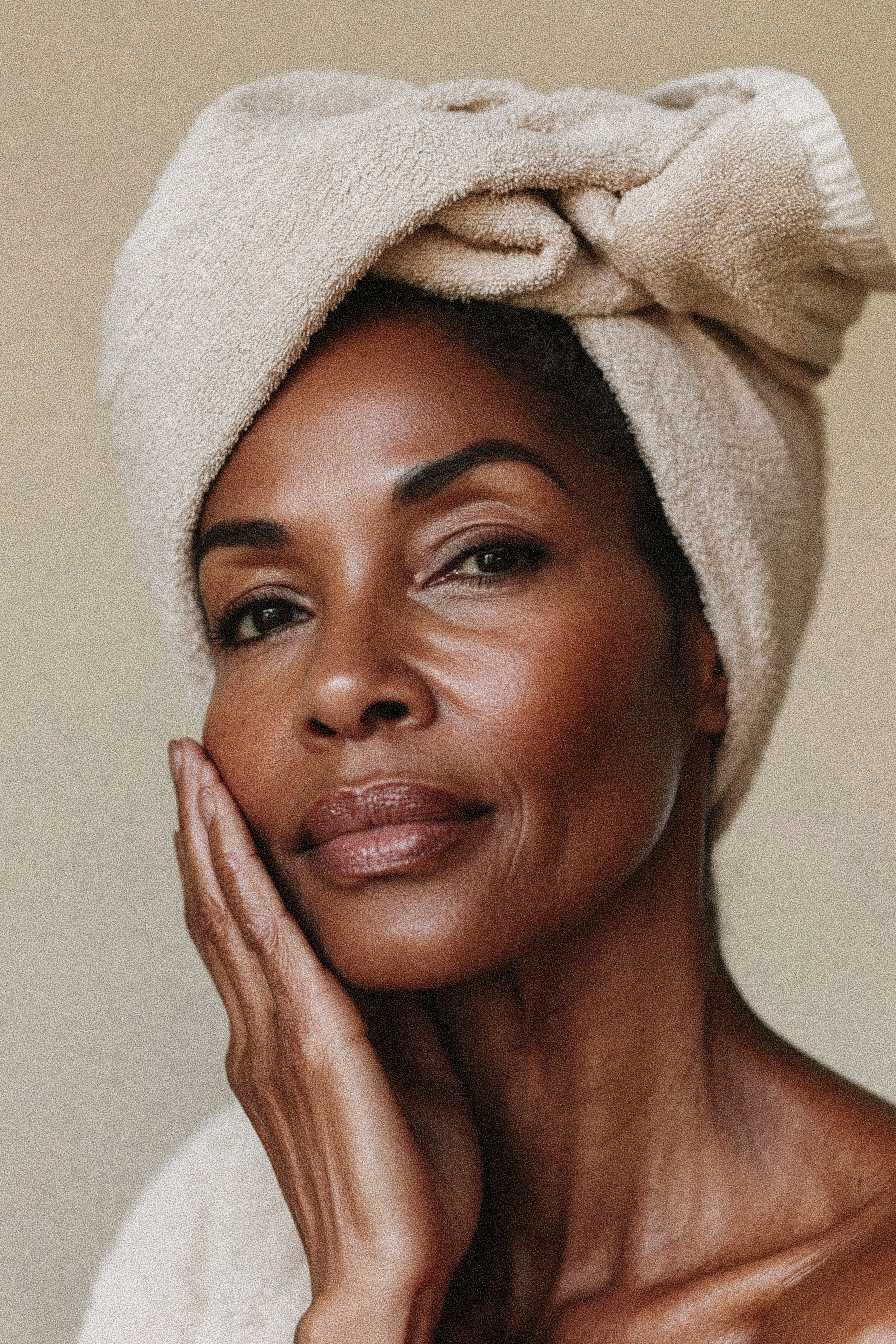 A woman with dark skin and short hair, wearing a beige towel wrapped around her head, touching her face gently with her hand.