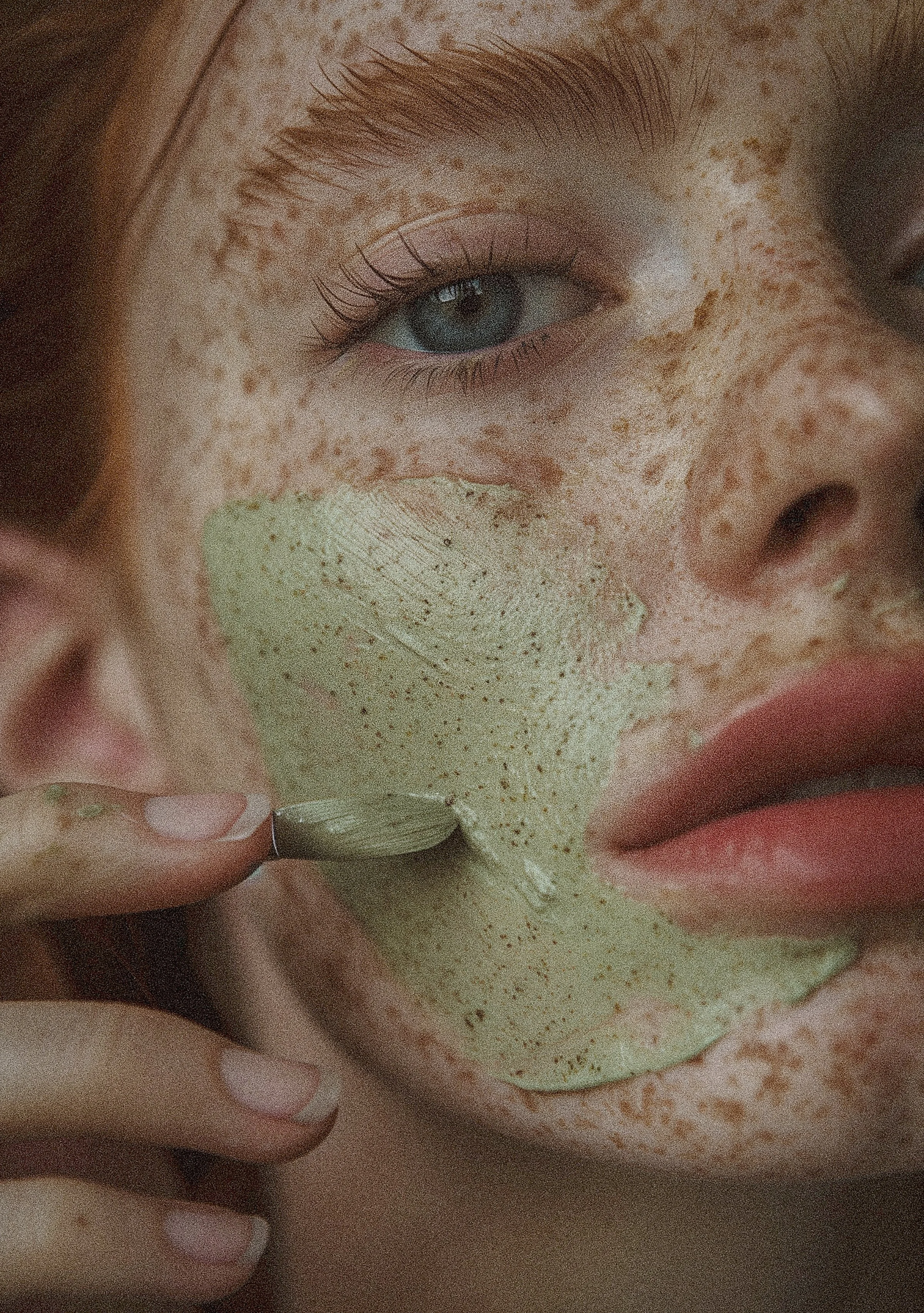 A close-up of a person's face showing the eye, nose, lips, and freckles, with fingers touching the face and a clay mask applied.