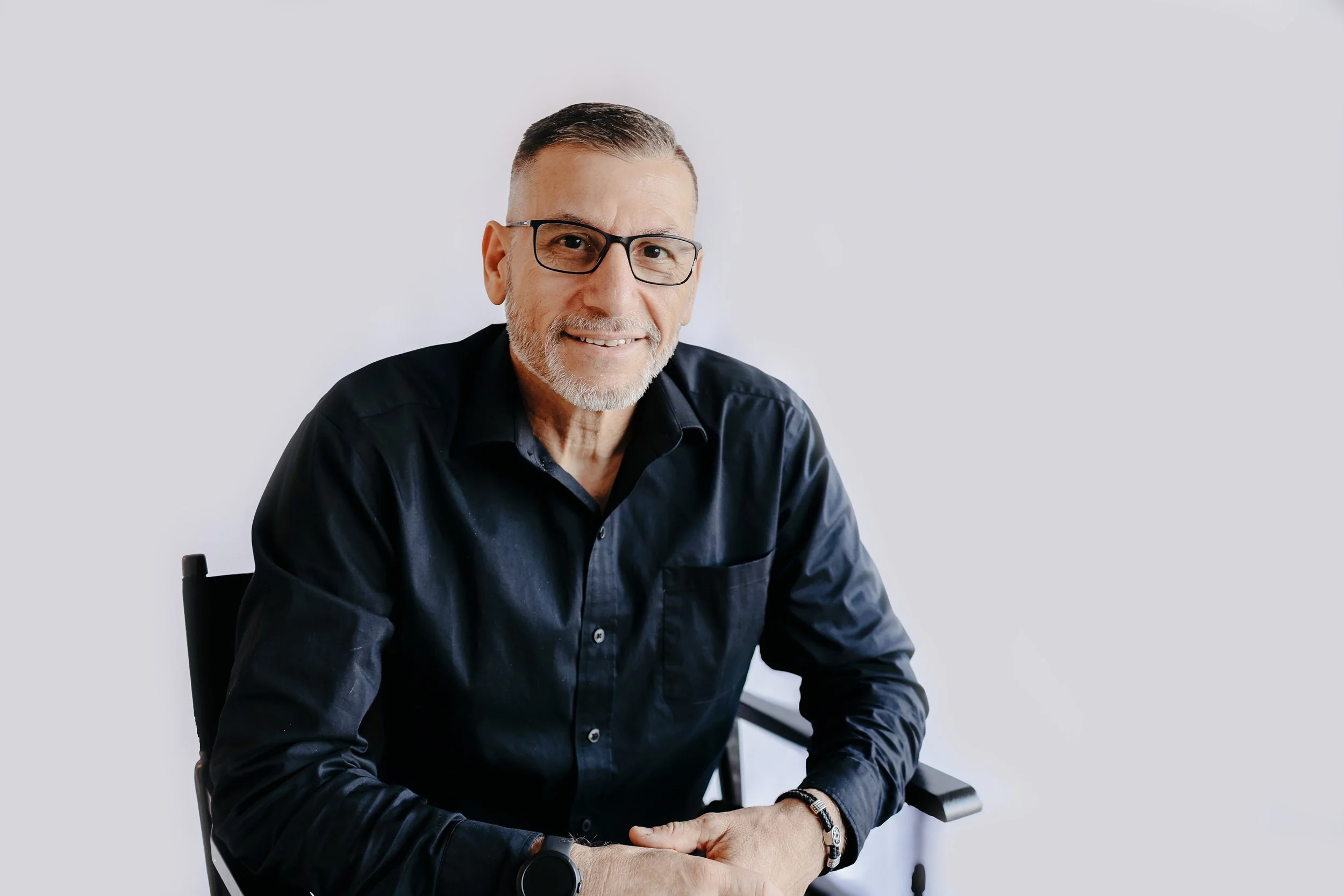 A smiling middle-aged man with gray hair and beard, wearing black glasses and a black shirt, sitting in a wheelchair against a plain white background.