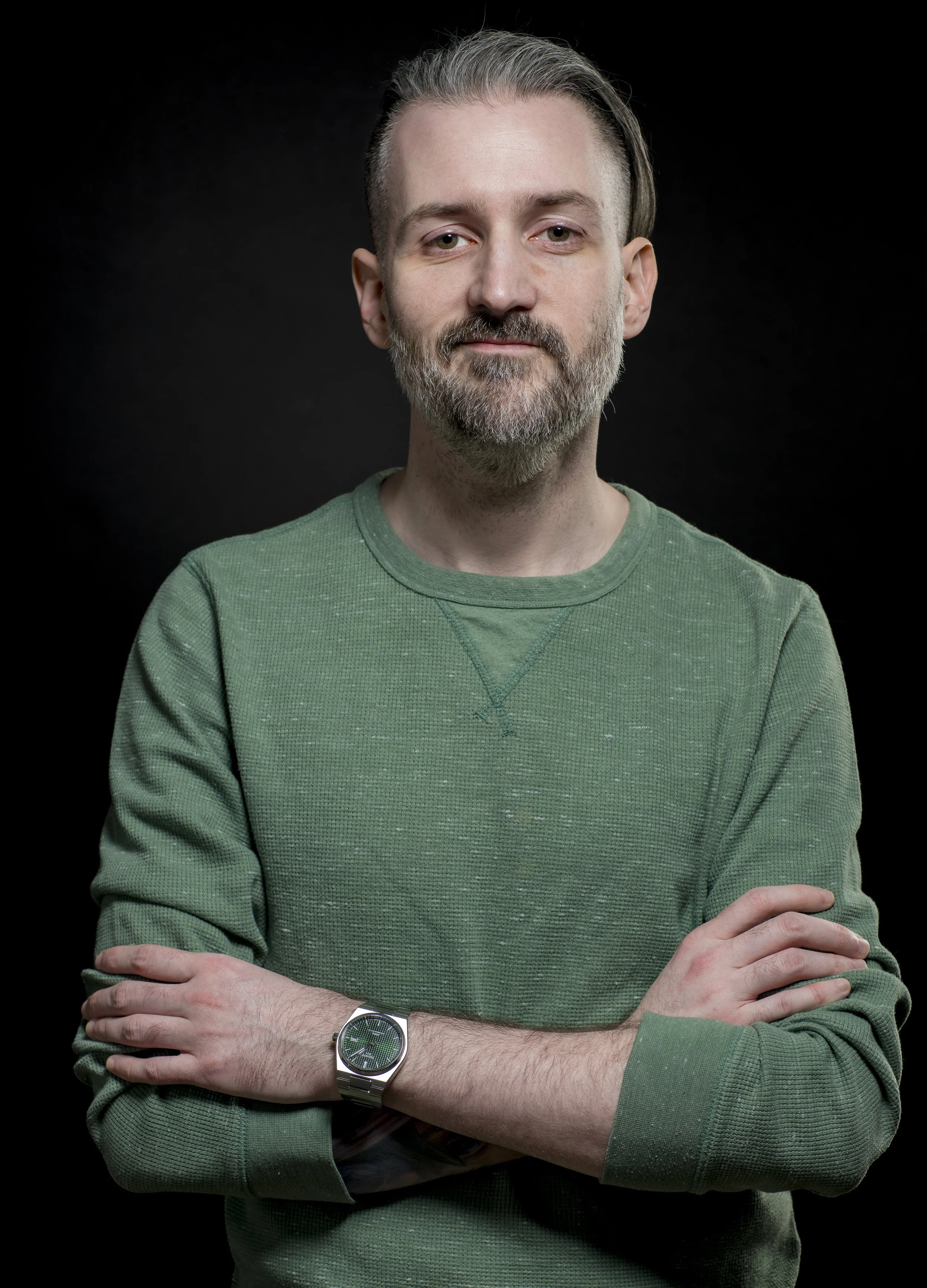 A man with gray hair and beard wearing a green sweater and silver watch, standing against a black background.