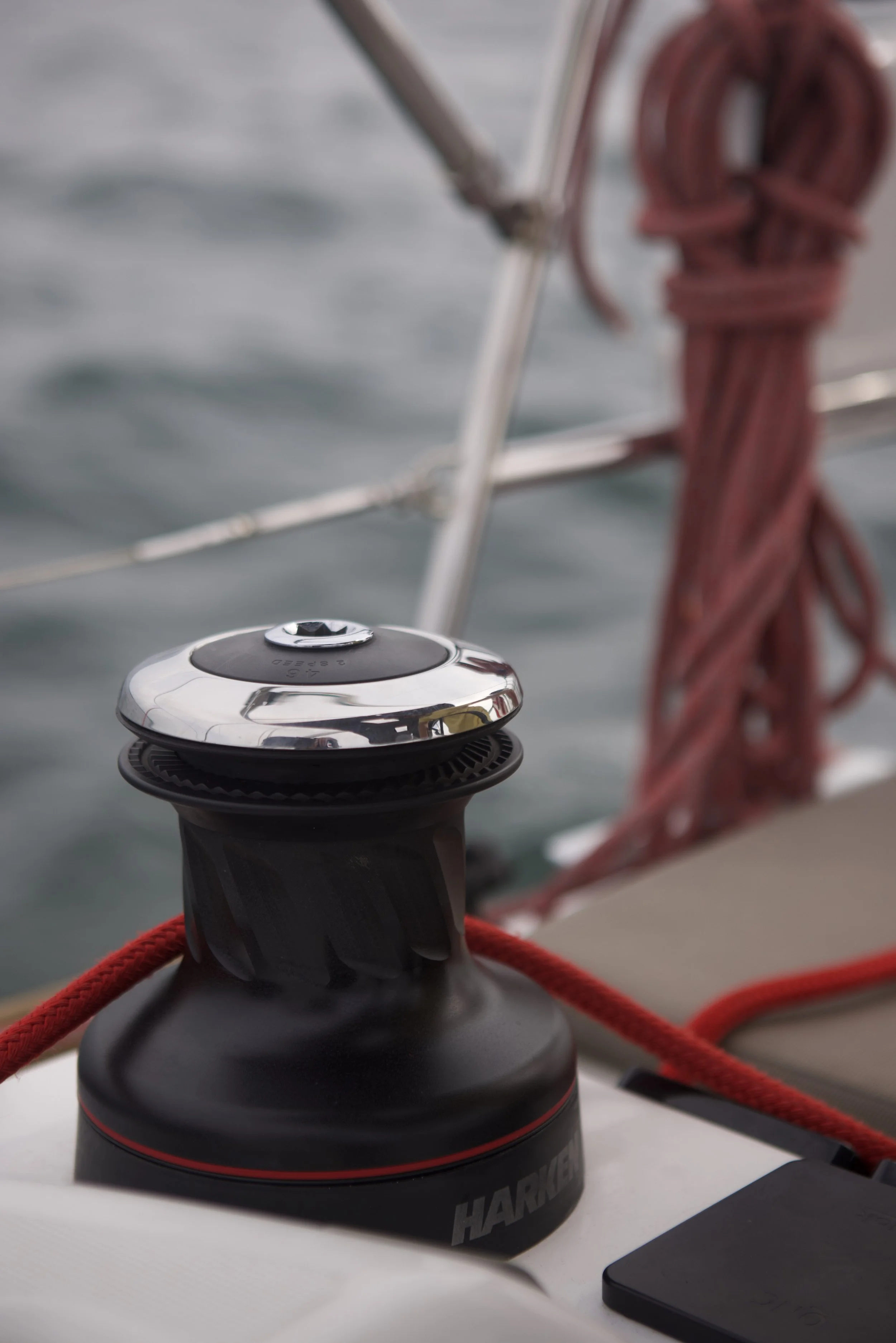 Close-up of a black and silver boat winch with red ropes tied around it on a boat with blurred water and red coil rope in the background.