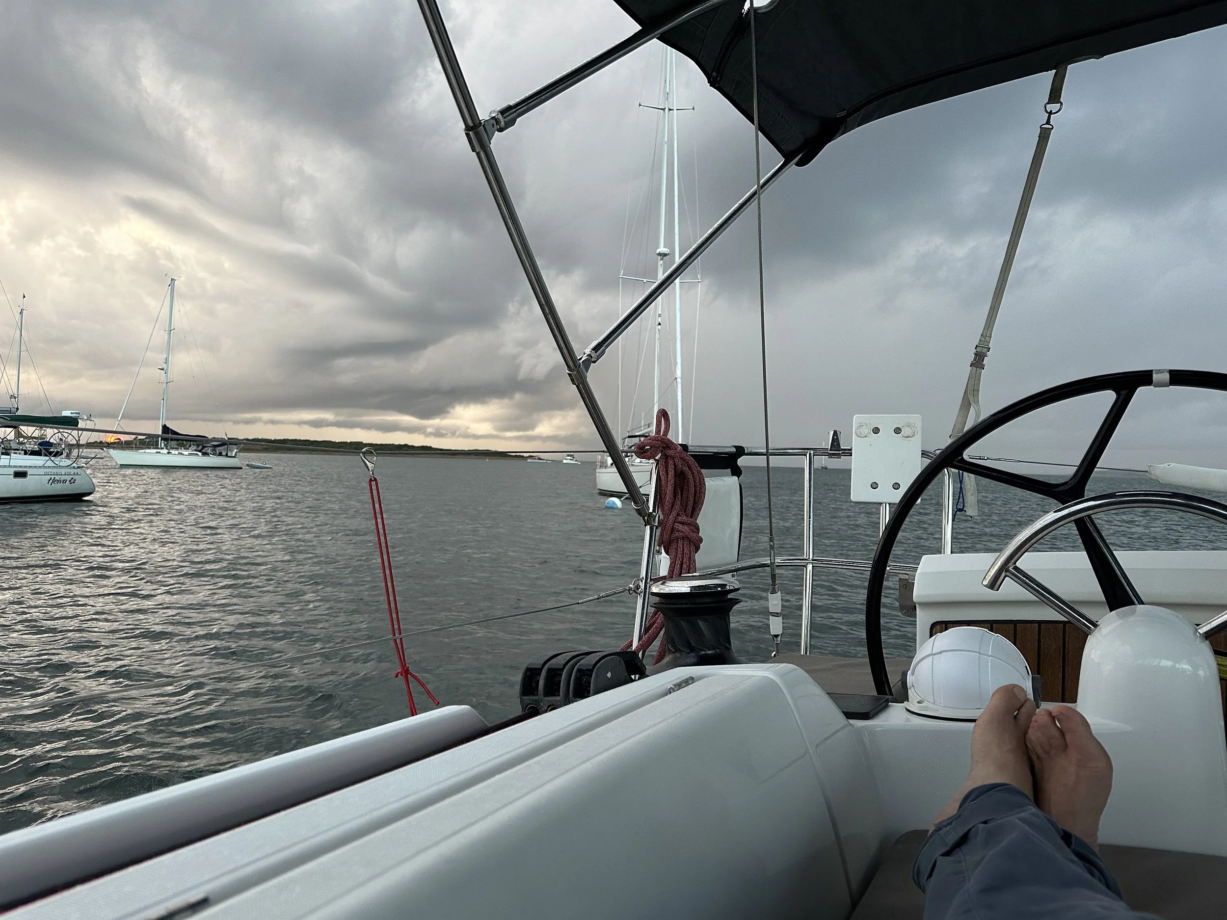 A view from a boat with gray clouds overhead, showing the boat's steering wheel, a person's feet, and other sailboats in the water nearby.
