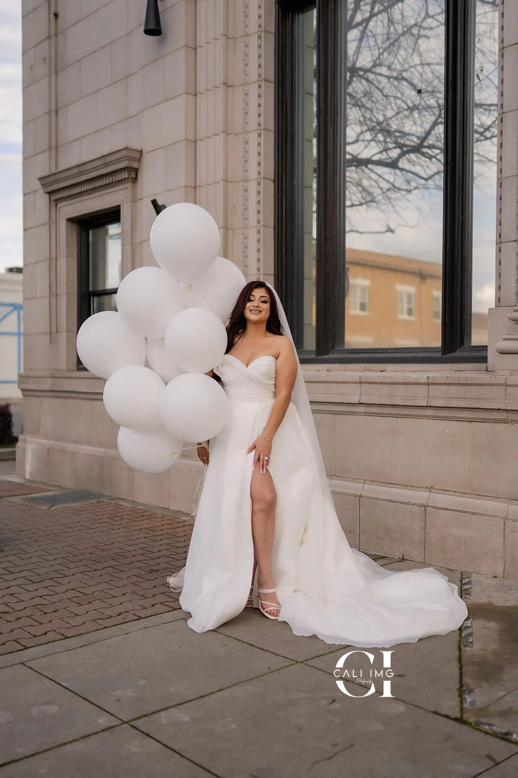 A smiling bride in a strapless white wedding gown with a slit, standing outside in front of a building with large windows, holding a large bunch of white balloons, with brick and sidewalk visible in the surroundings.