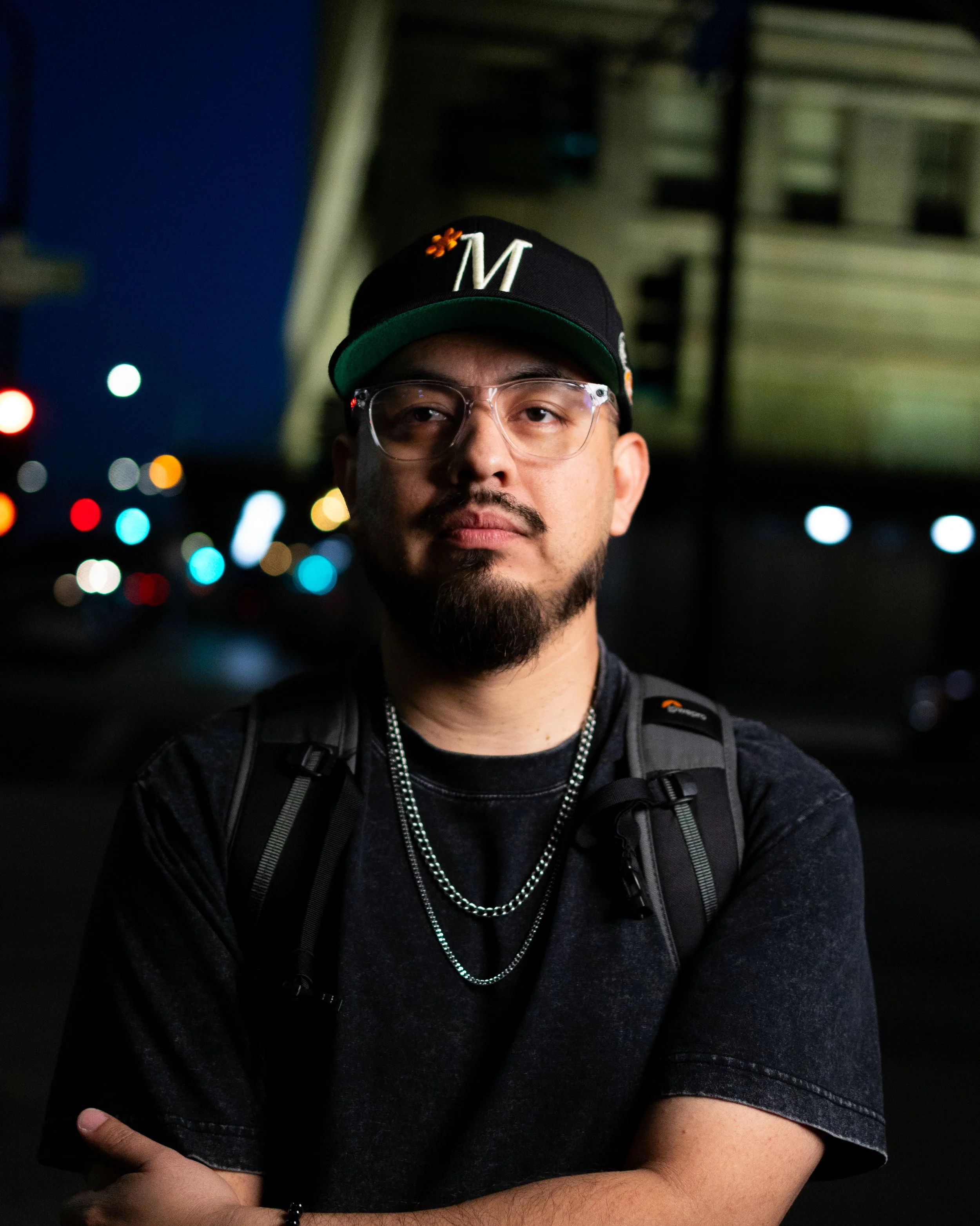 A young man with glasses, a goatee, and a beard standing outdoors at night, wearing a black cap with a white 'M' and orange flower, a black backpack, and chains, with city lights blurred in the background.