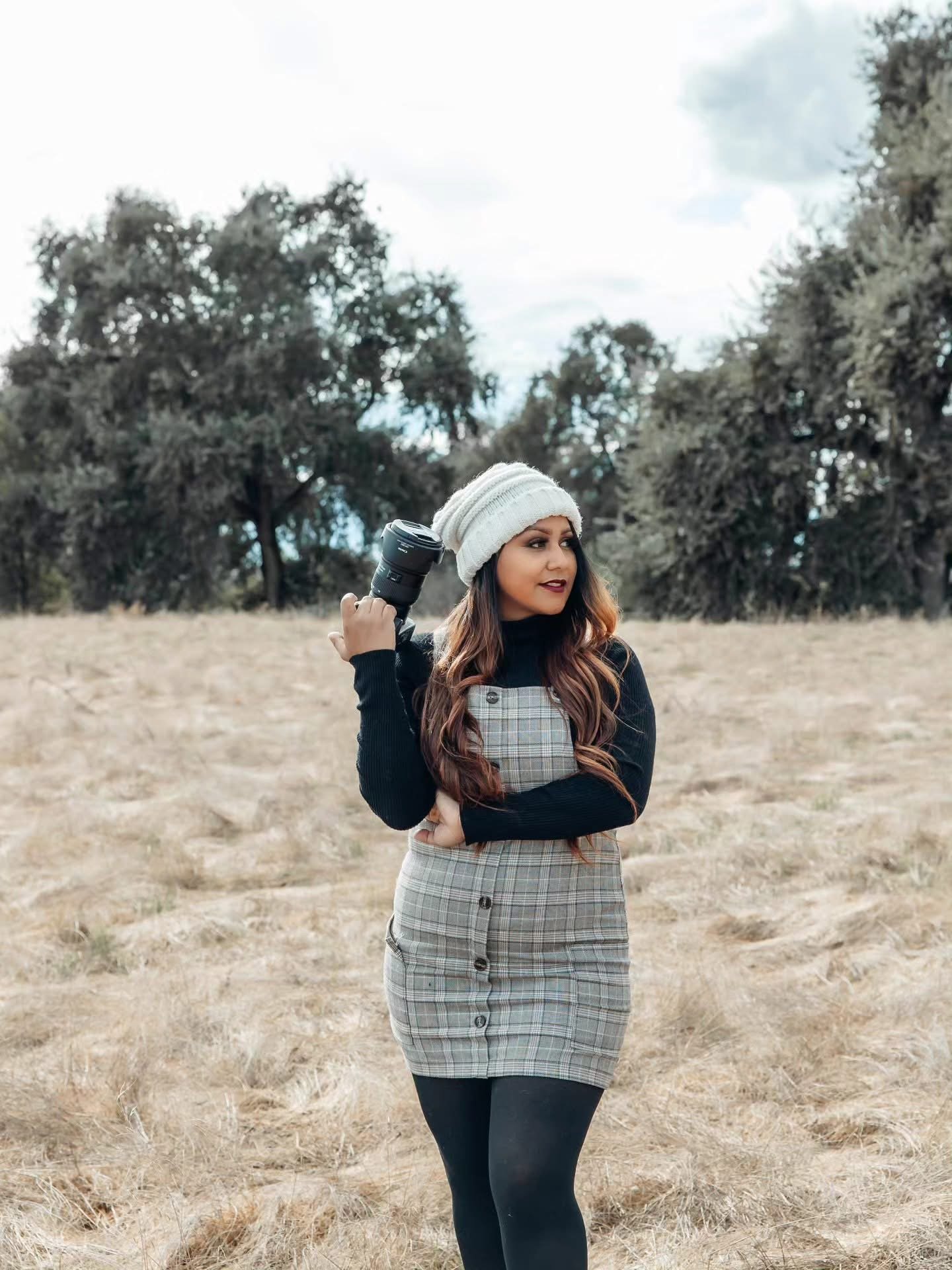 A woman standing outdoors in a field with trees in the background, holding a camera on her shoulder, wearing a white knit beanie, black top, plaid dress, and black tights.