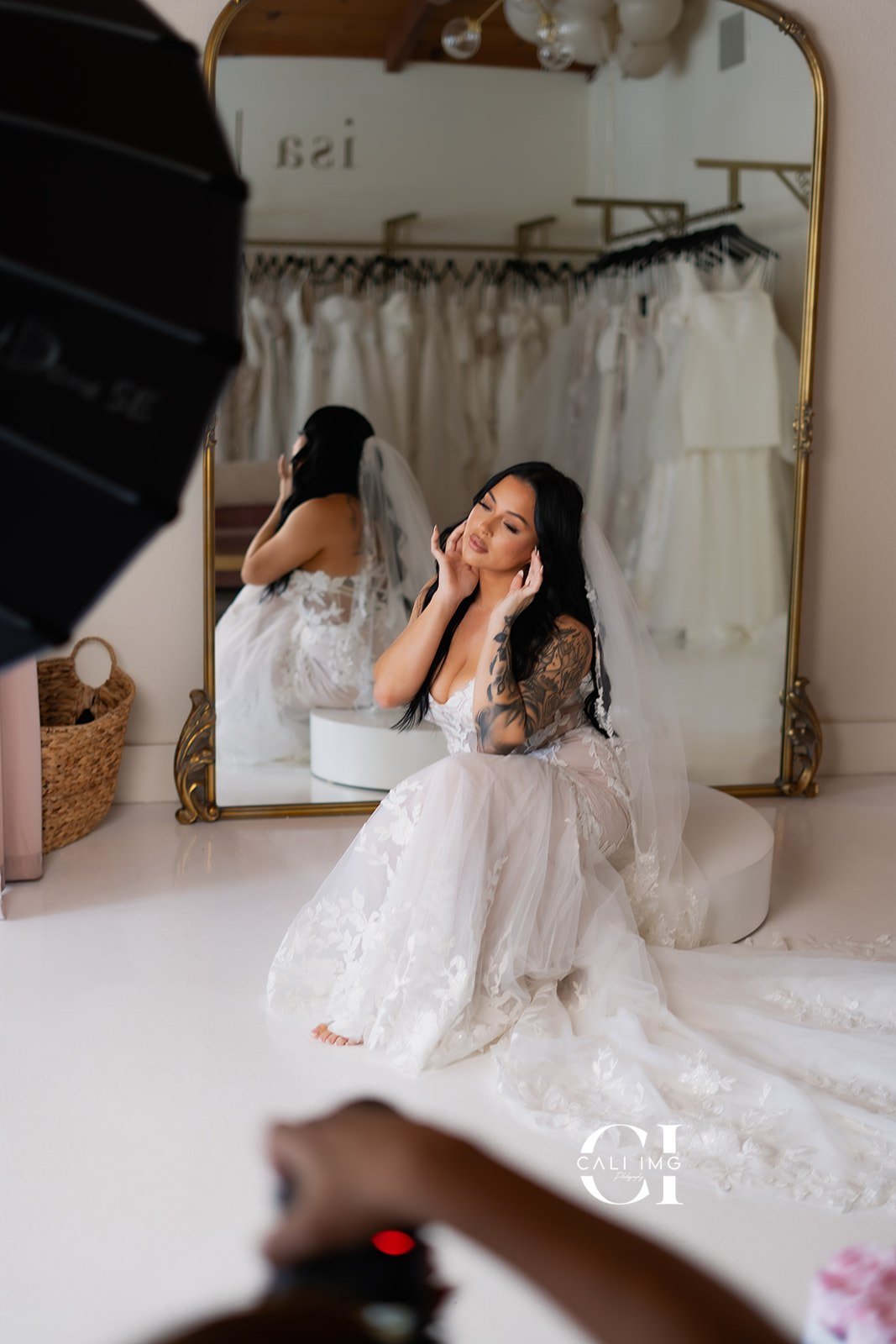 A bride with long dark hair and tattoos on her arms, wearing a white wedding dress and veil, sits on a white surface in front of a large mirror. She is gently touching her face with her eyes closed, surrounded by wedding dresses hanging in the background.