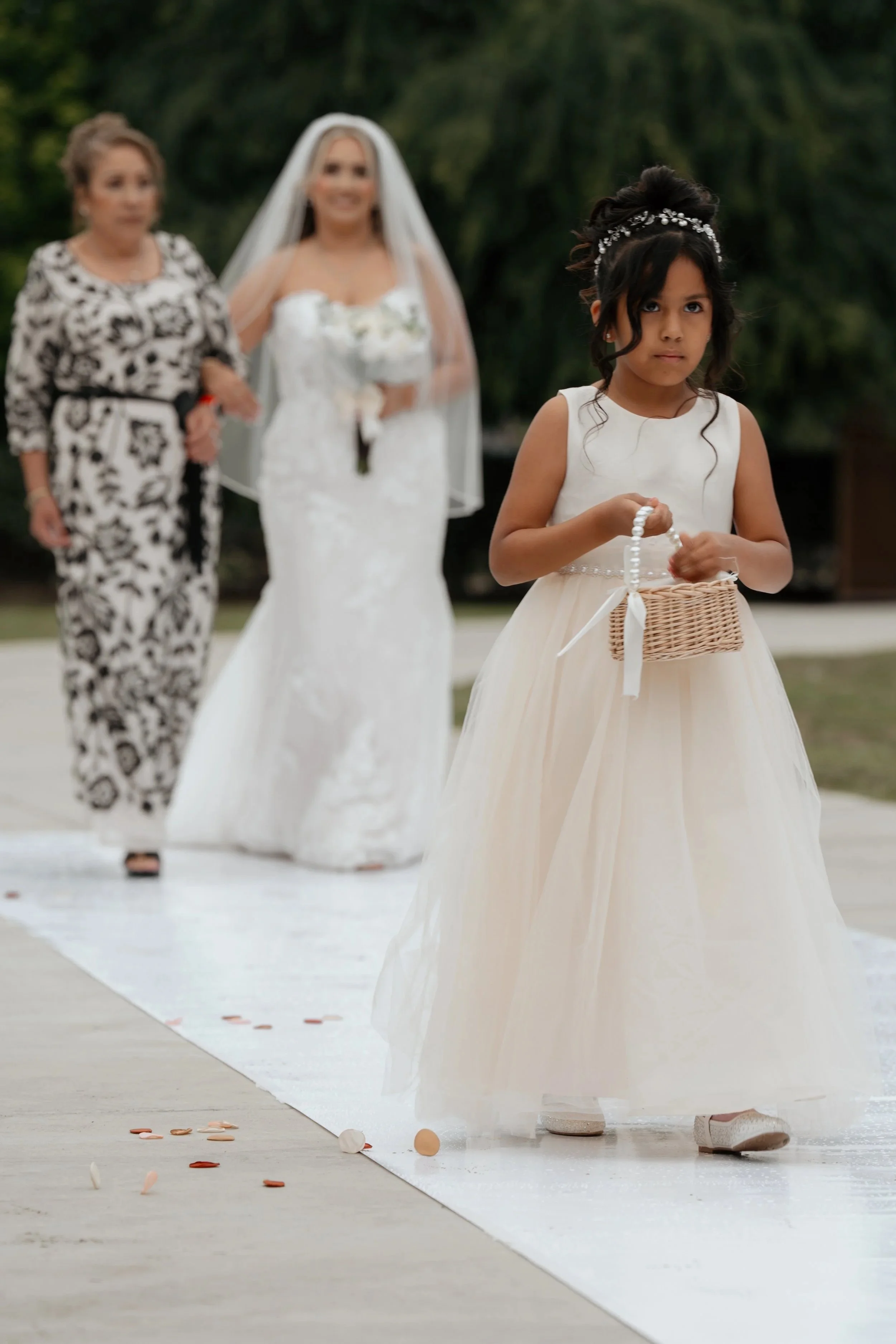 Young girl in a white gown with a pearl headband holding a basket, standing on a white aisle with colored rose petals, during a wedding ceremony outdoors. Blurred woman in a floral dress and a bride in a white wedding dress in the background.