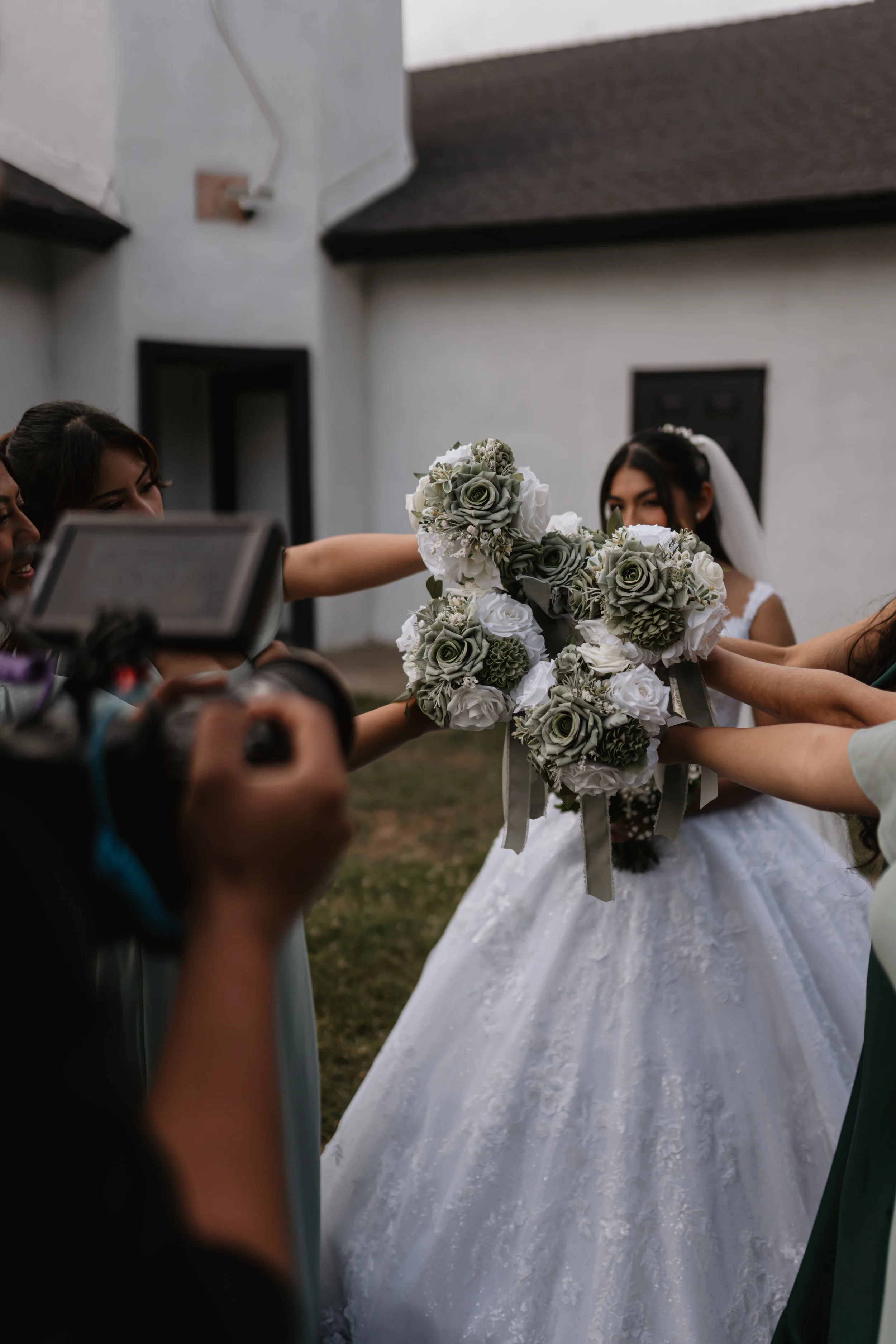 A bride holding a bouquet of white and gray flowers among friends or bridesmaids during a wedding celebration.