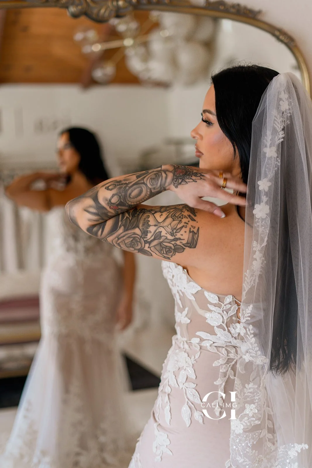A bride adjusting her wedding dress with an intricate tattoo sleeve on her right arm, standing in front of a mirror while another bride in a lace wedding gown is seen in the background.